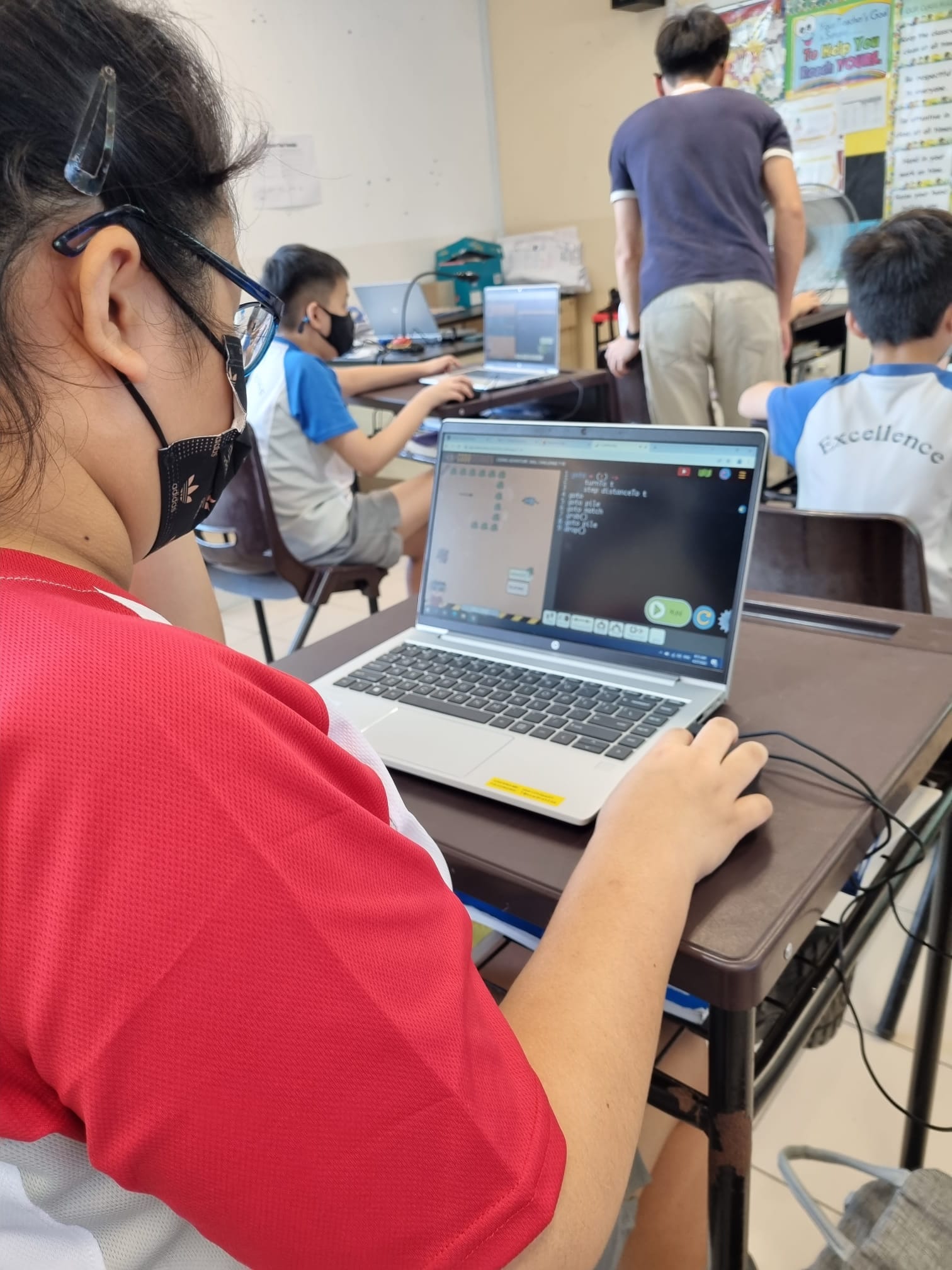 Student wearing a mask and glasses uses a laptop with a coding program in a classroom with other students.