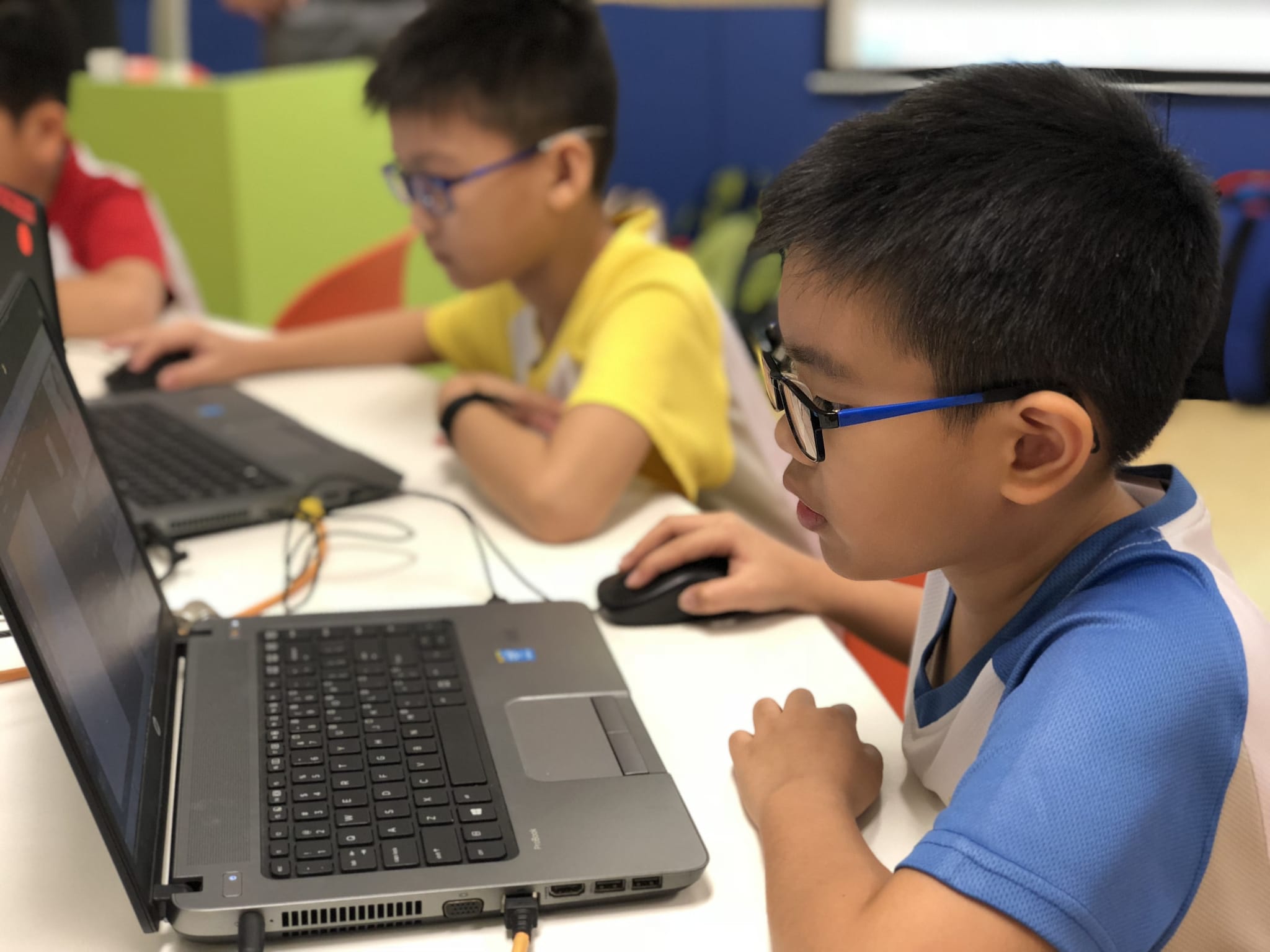 Two young boys wearing glasses work on laptops at a white desk in a colorful classroom.