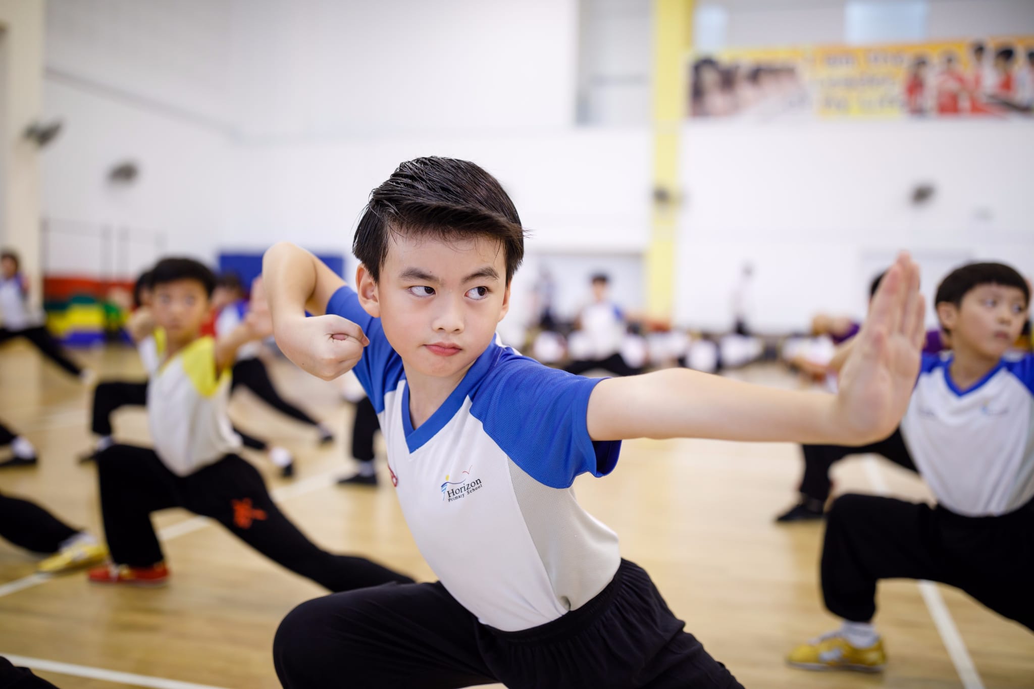A boy in a blue and white school shirt performs a martial arts stance in a gym with other children behind him.