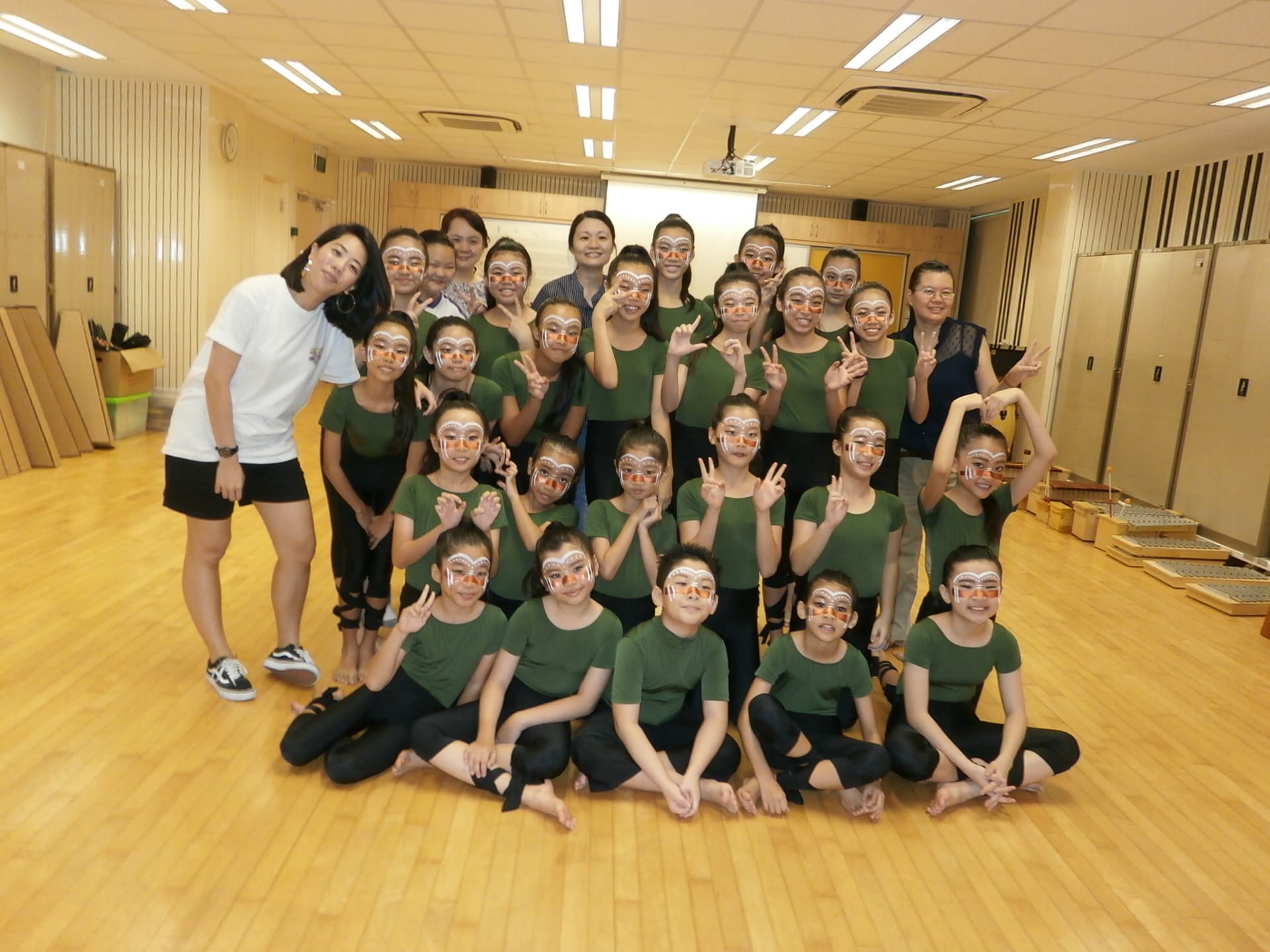 Group of children in green outfits with orange and white face paint pose with adults in a dance studio.