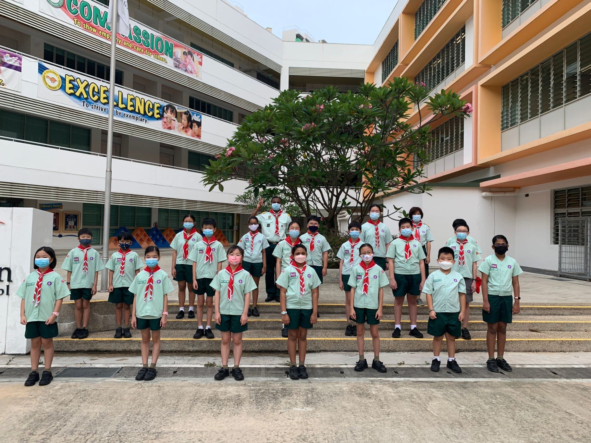 A group of children in green scout uniforms with red neckerchiefs and face masks pose outdoors at a school.