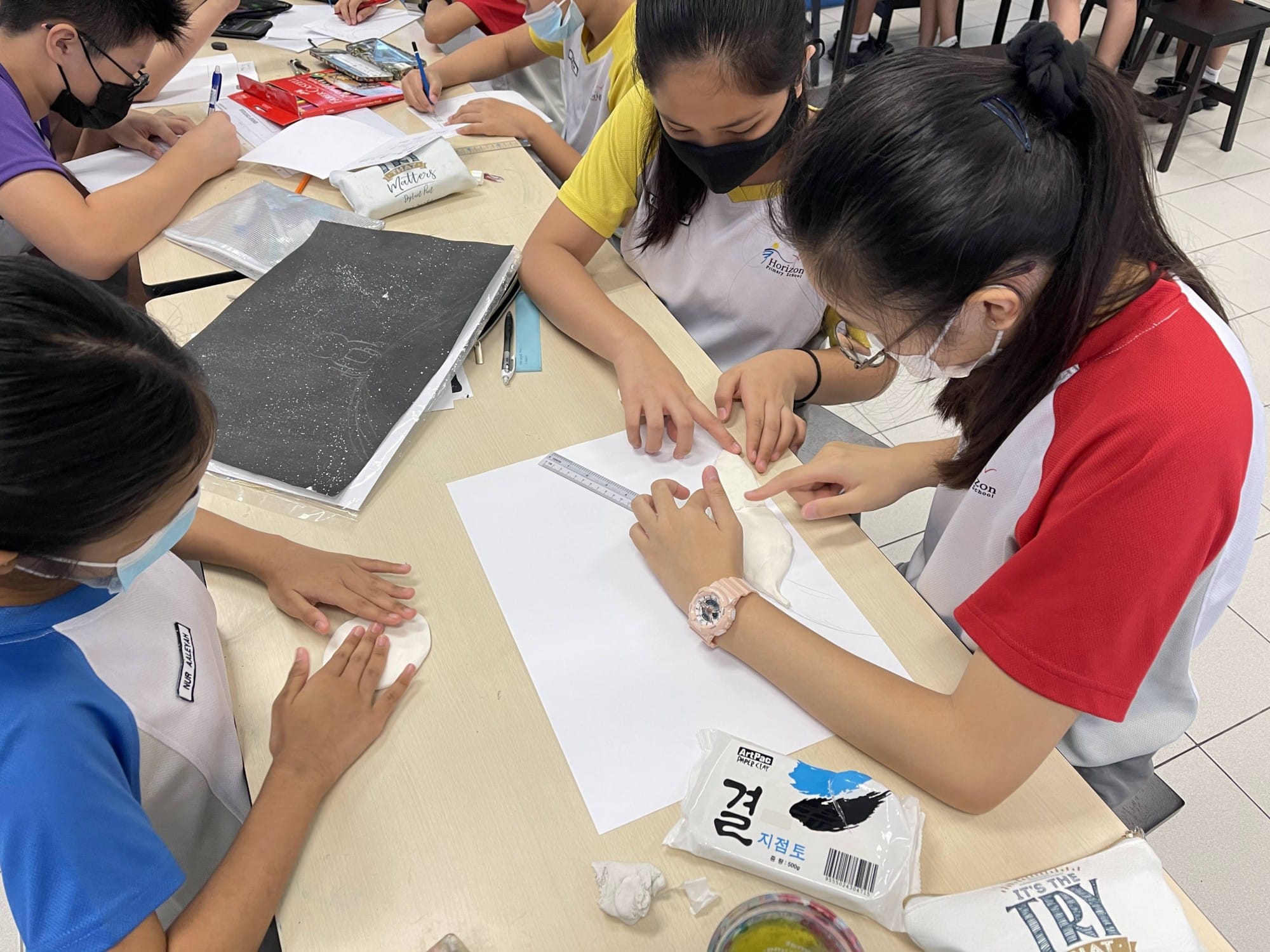 Students wearing masks work together at a table, shaping clay and using a ruler on white paper.