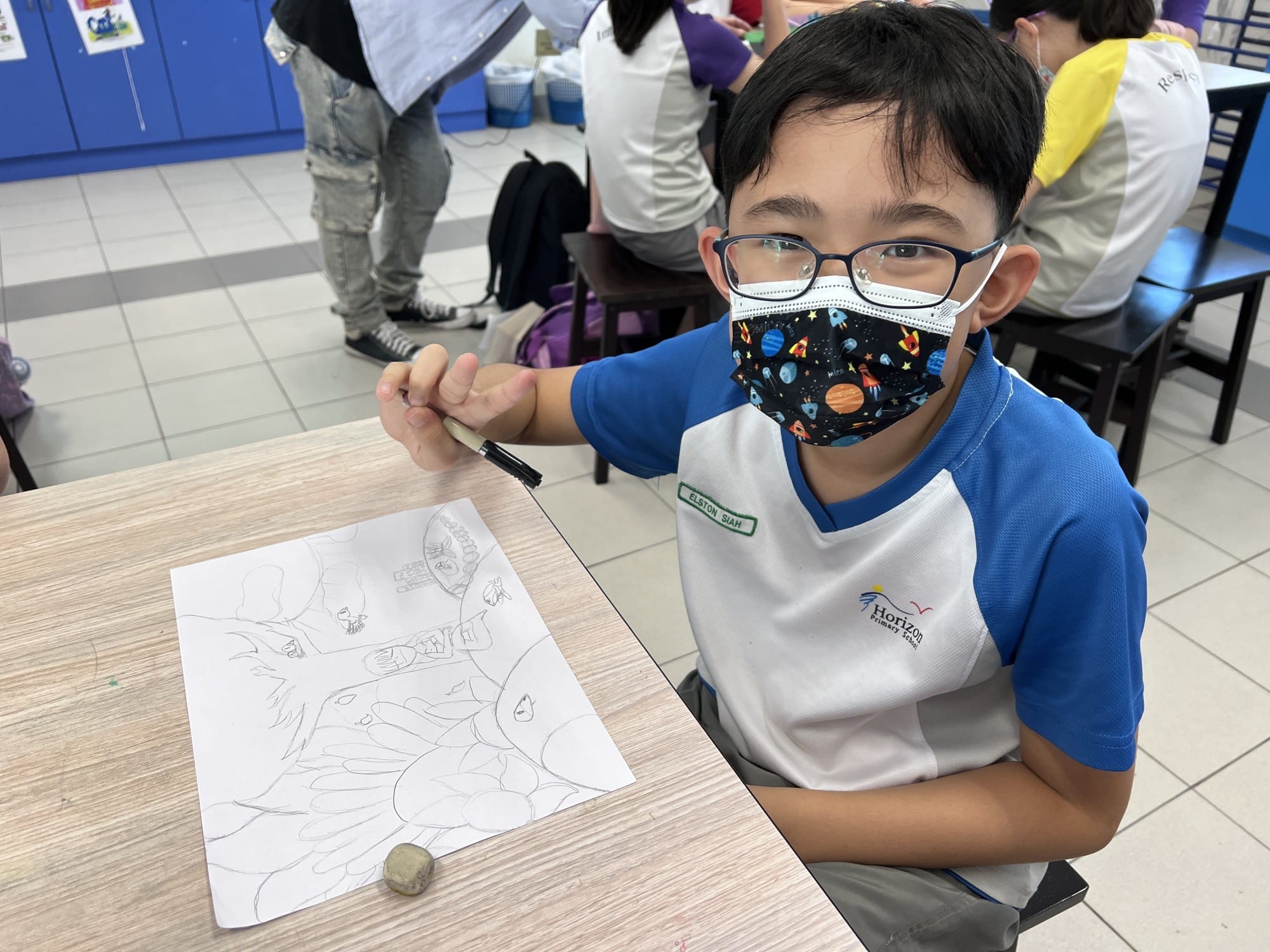 A masked boy wearing glasses sits at a desk holding a pen beside his detailed pencil sketch in a classroom.