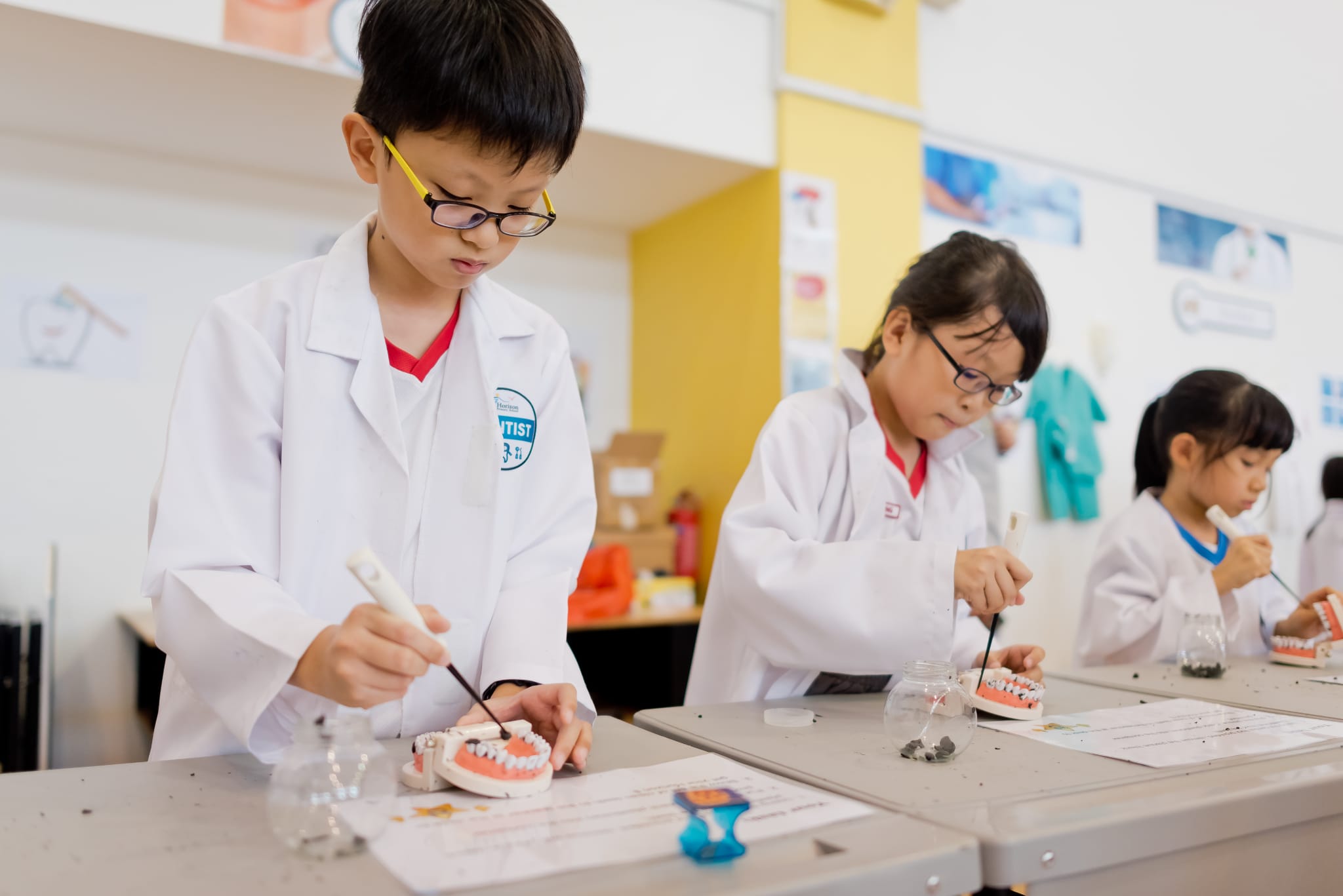 Three children in white lab coats use small tools on dental jaw models at classroom desks.