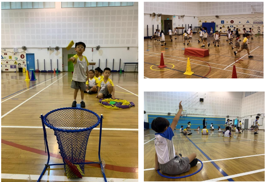 Young children in a school gymnasium doing physical activities with cones, hoops, and a basket net.