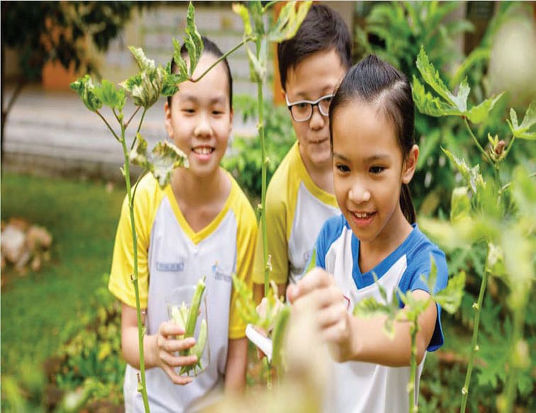 Three smiling children in school uniforms examine green plants outdoors, with one girl touching a stem.