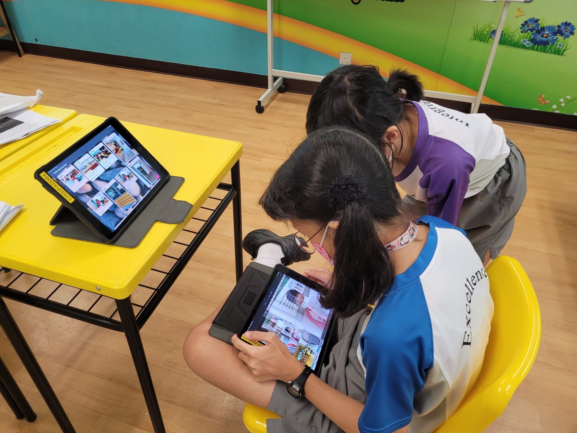 Two students in school uniforms lean over tablets in a colorful classroom with yellow desks.