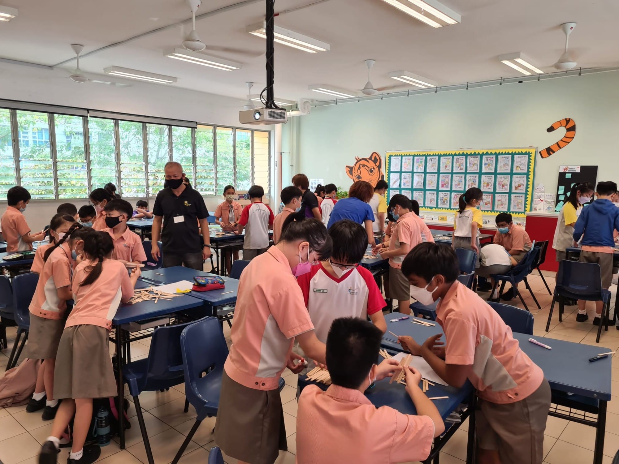 Students in pink uniforms work with craft sticks at blue desks in a bright classroom supervised by masked adults.