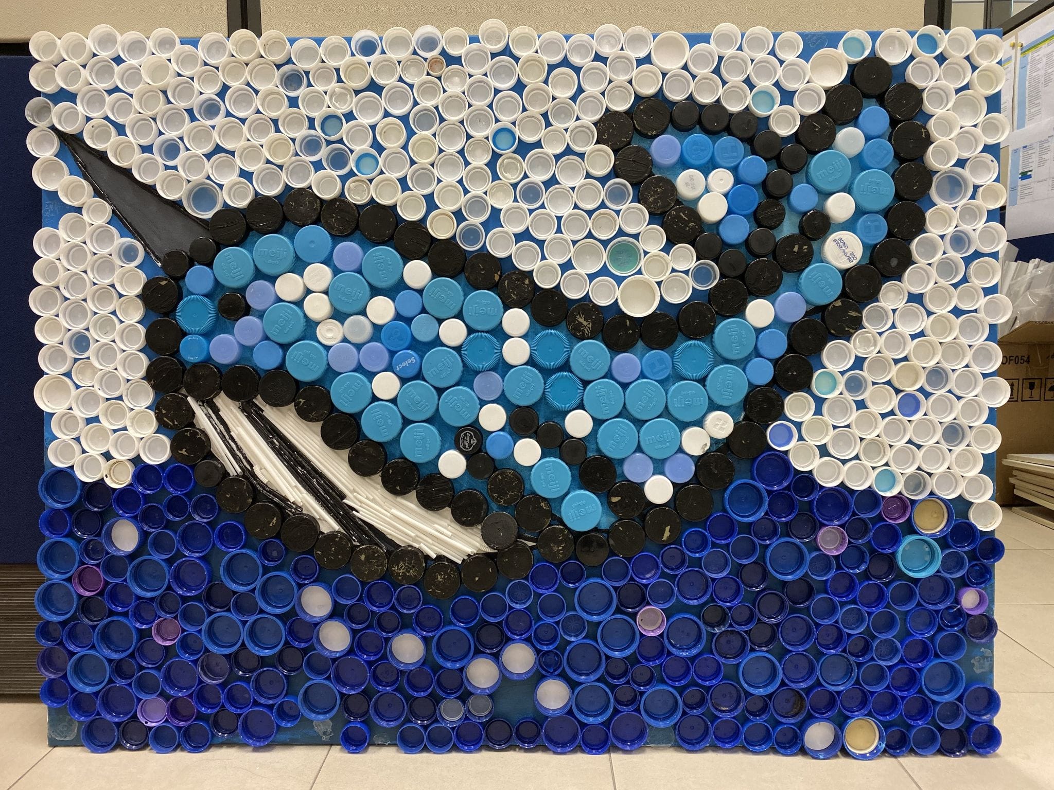 Mosaic artwork of an orca whale made from hundreds of blue, white, and black plastic bottle caps.