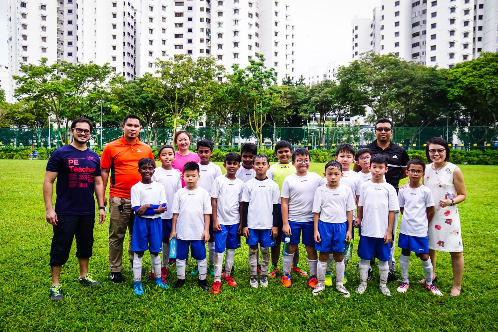 A youth soccer team in white shirts and blue shorts poses with four adult coaches on a grass field near tall buildings.