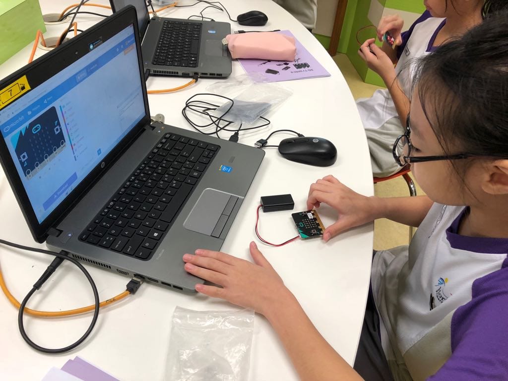 A young girl with glasses works on a micro:bit circuit board next to an HP laptop showing a coding interface.