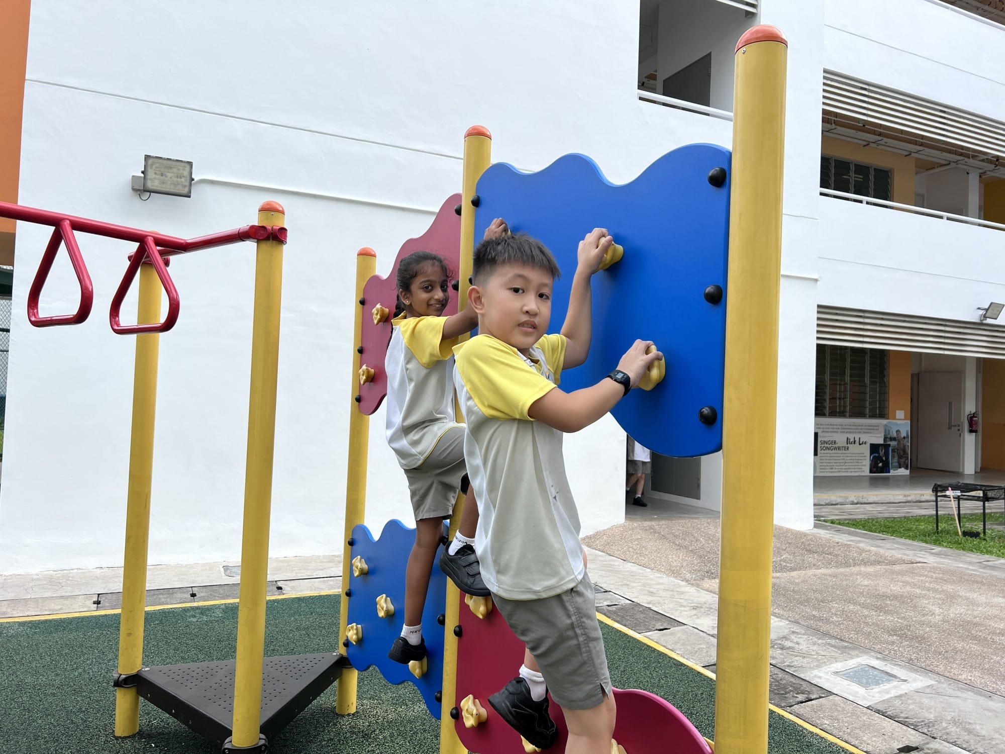 Two children in school uniforms climb a colorful blue and red playground structure with yellow poles.