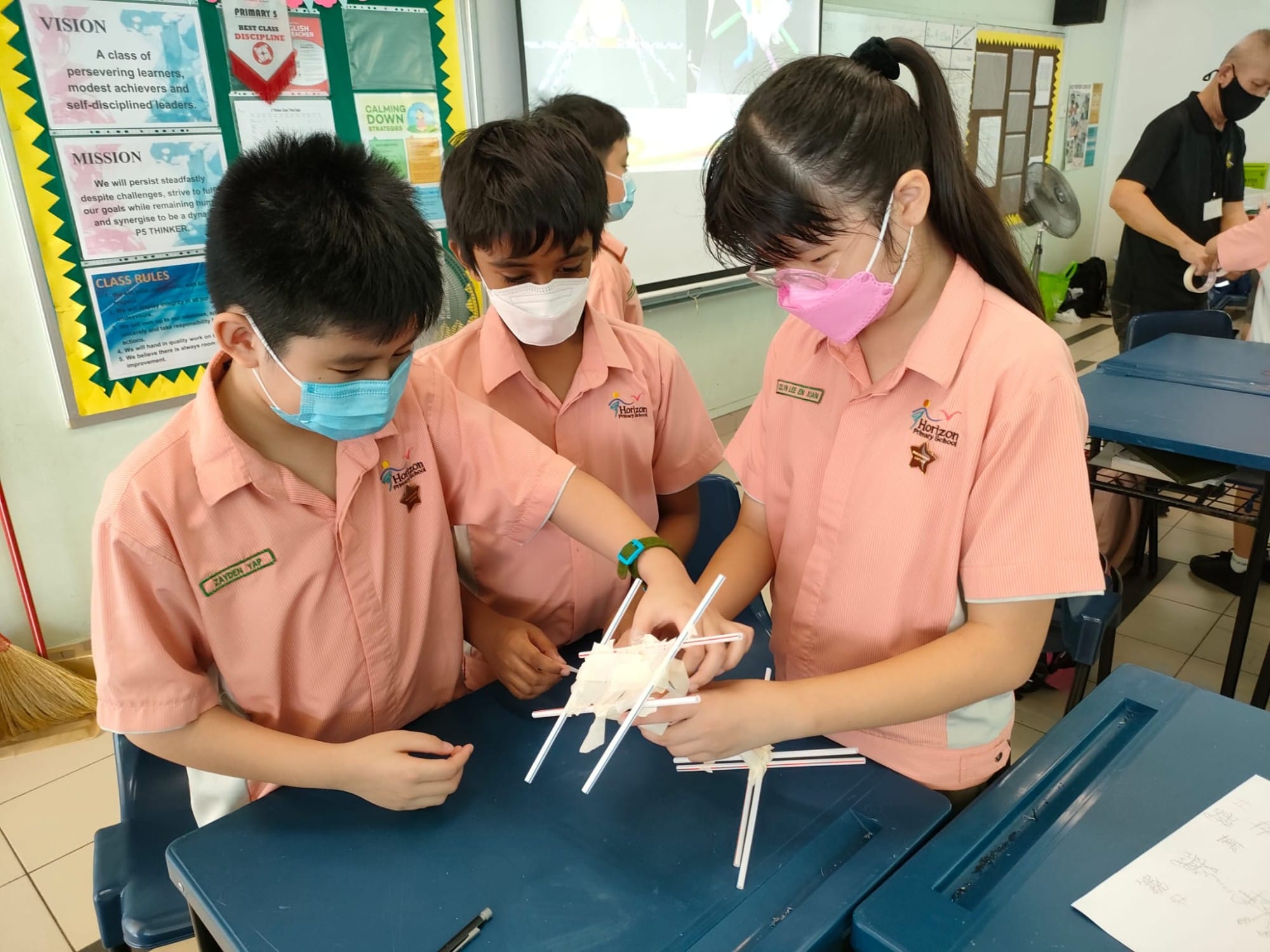 Three masked primary school students in pink uniforms collaborate on building a structure using straws at a classroom desk.