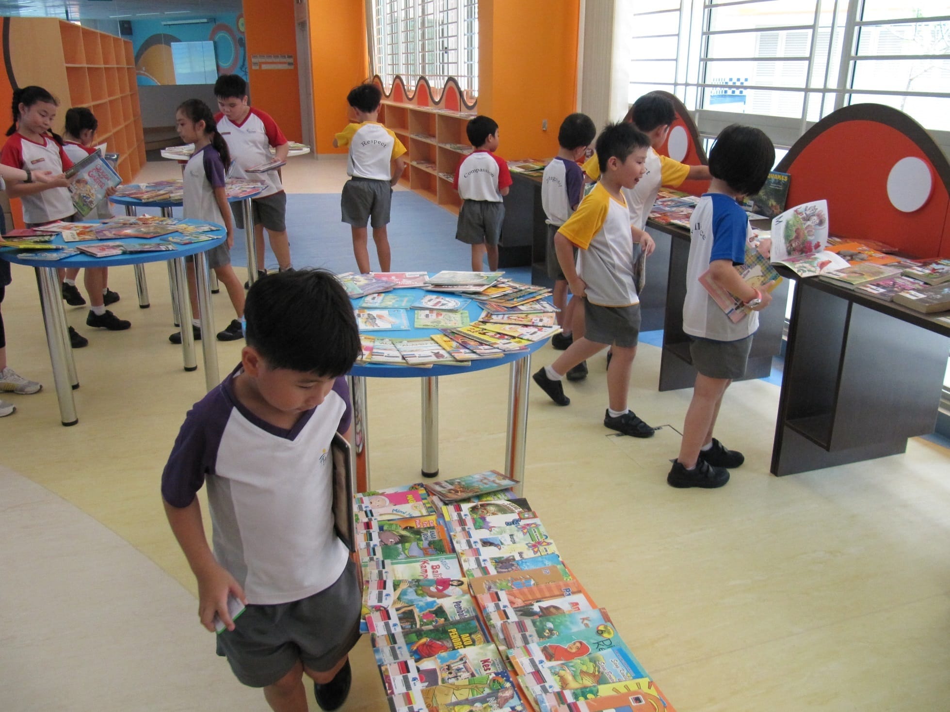 Children in school uniforms browse colorful books spread across round tables in a bright orange library.