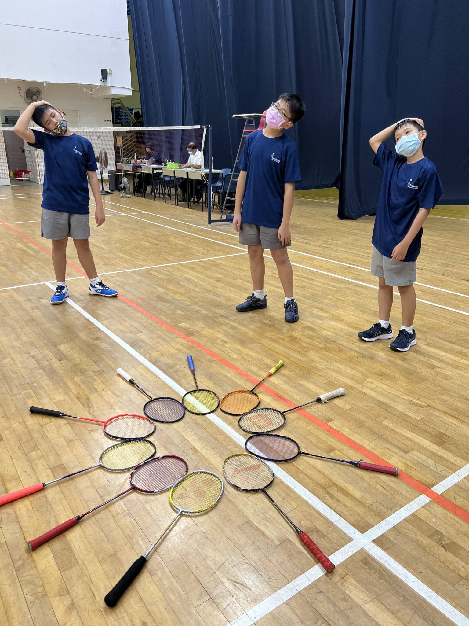 Three masked boys in blue shirts stand on a badminton court looking at many rackets arranged on the floor.