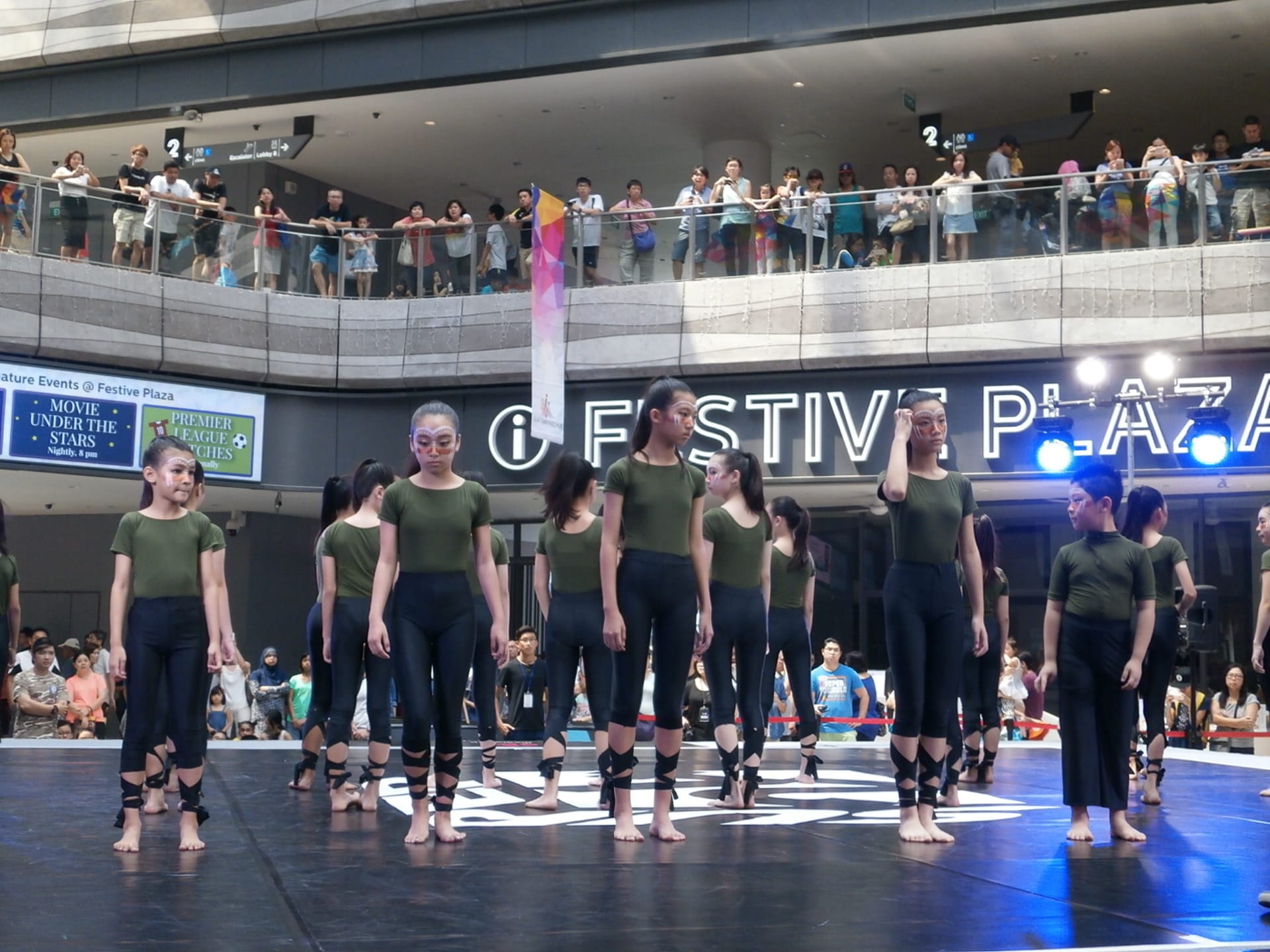 Young dancers in green tops and black leggings with face paint perform on a stage at Festive Plaza mall.