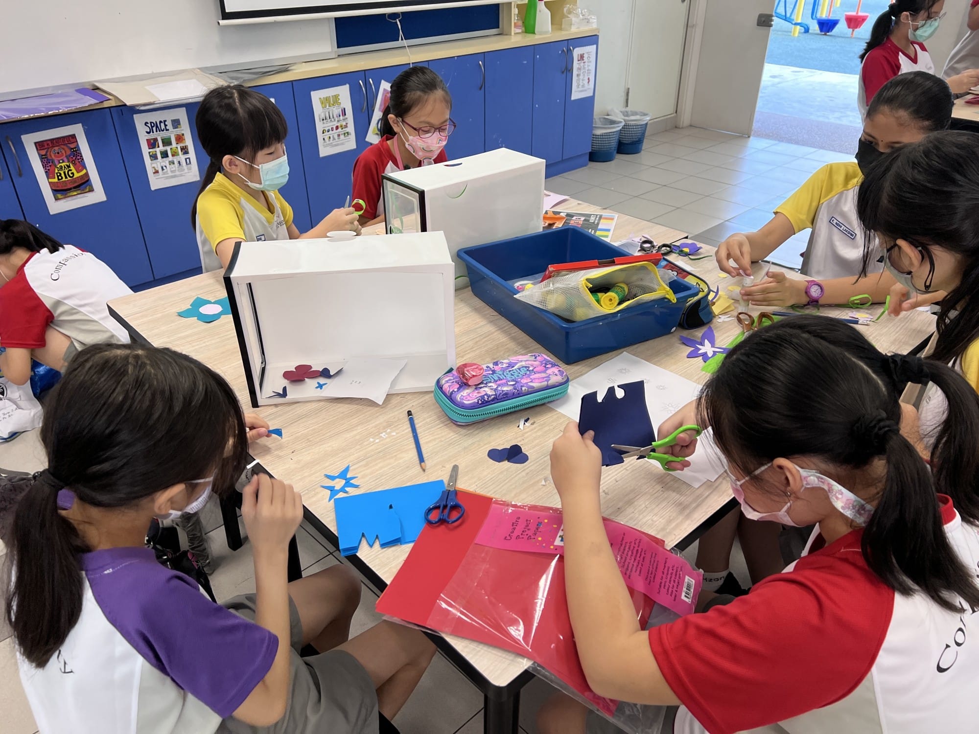 Children wearing masks sit around a classroom table cutting colorful paper shapes with scissors during an art activity.