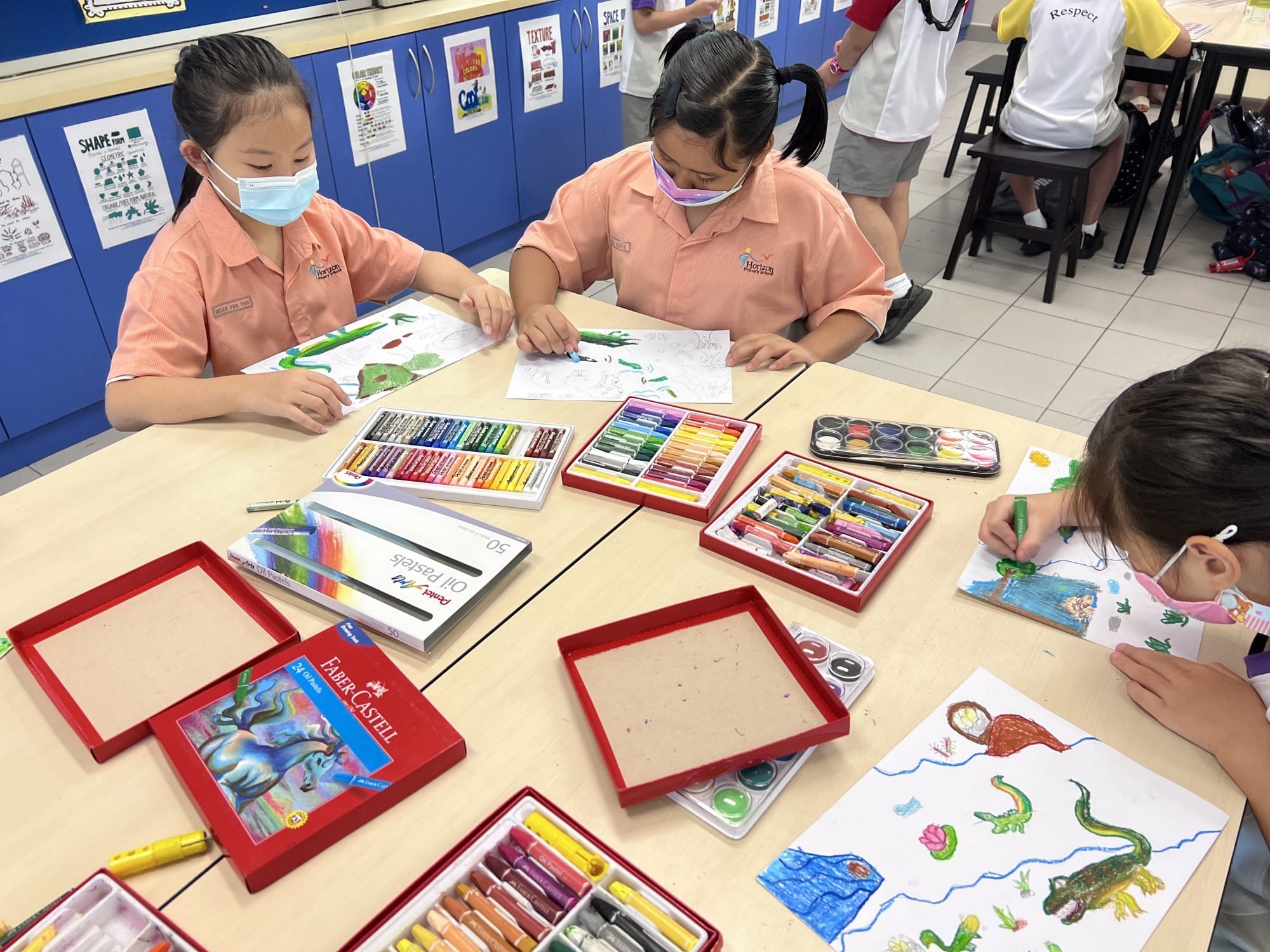 Three masked school children draw colorful dinosaur scenes at a table covered with oil pastels and watercolor sets.