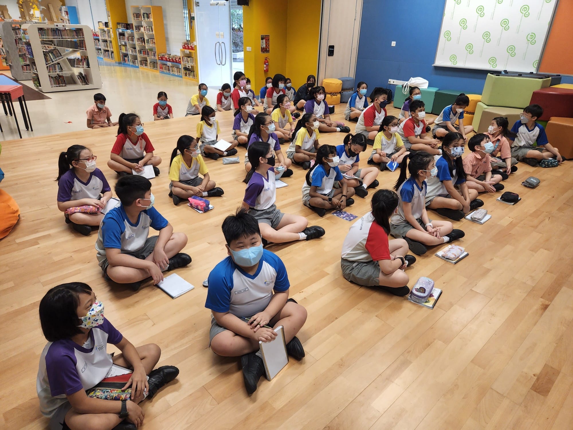 Students in colorful uniforms and face masks sit cross-legged on a wooden floor in a school library.
