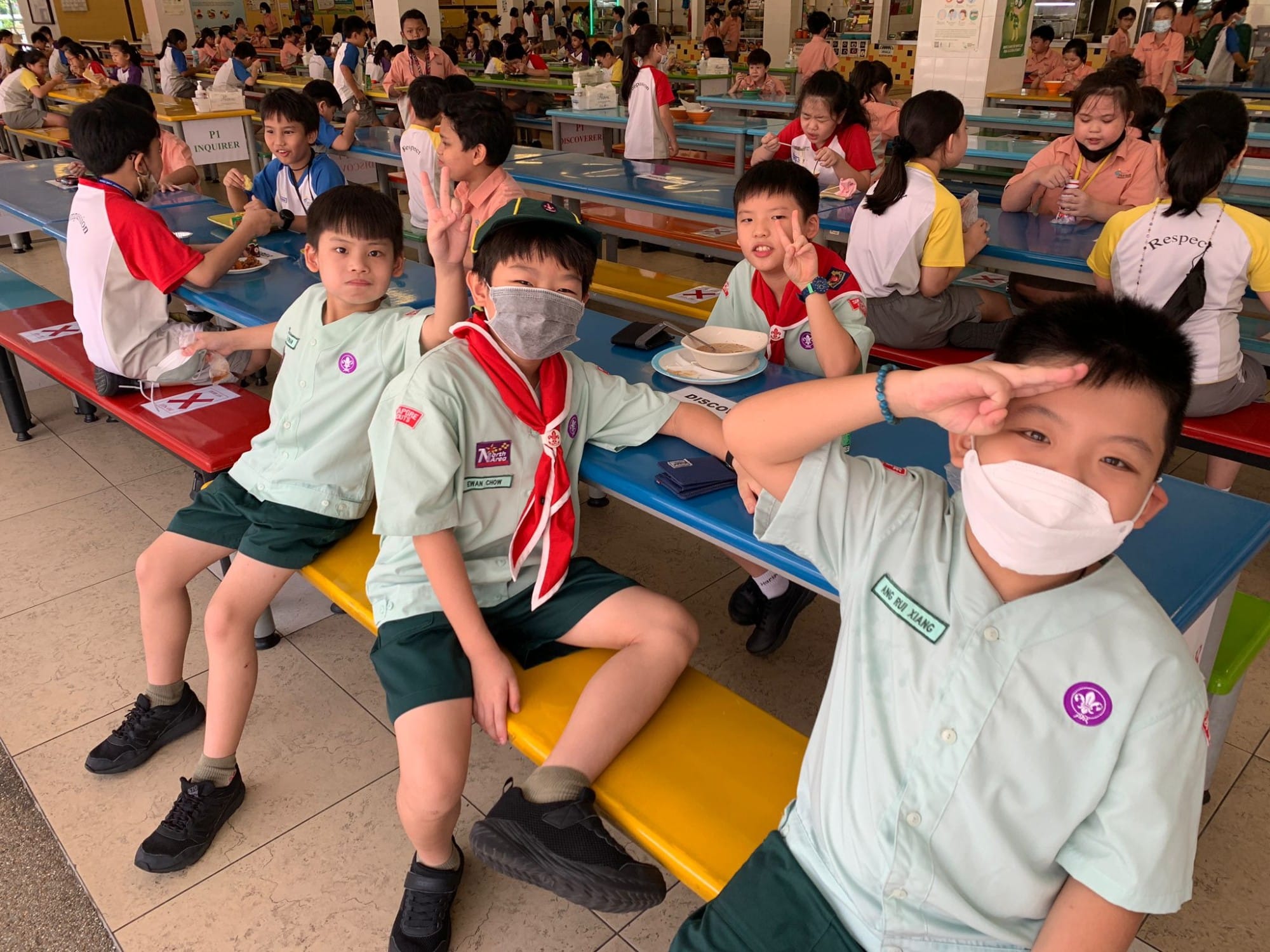 Young scouts in green uniforms and face masks pose at colorful school cafeteria benches filled with children eating.