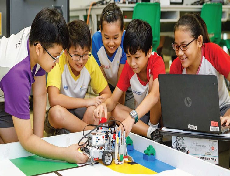 Five children gather around a LEGO Mindstorms robot and an HP laptop, smiling while working together.
