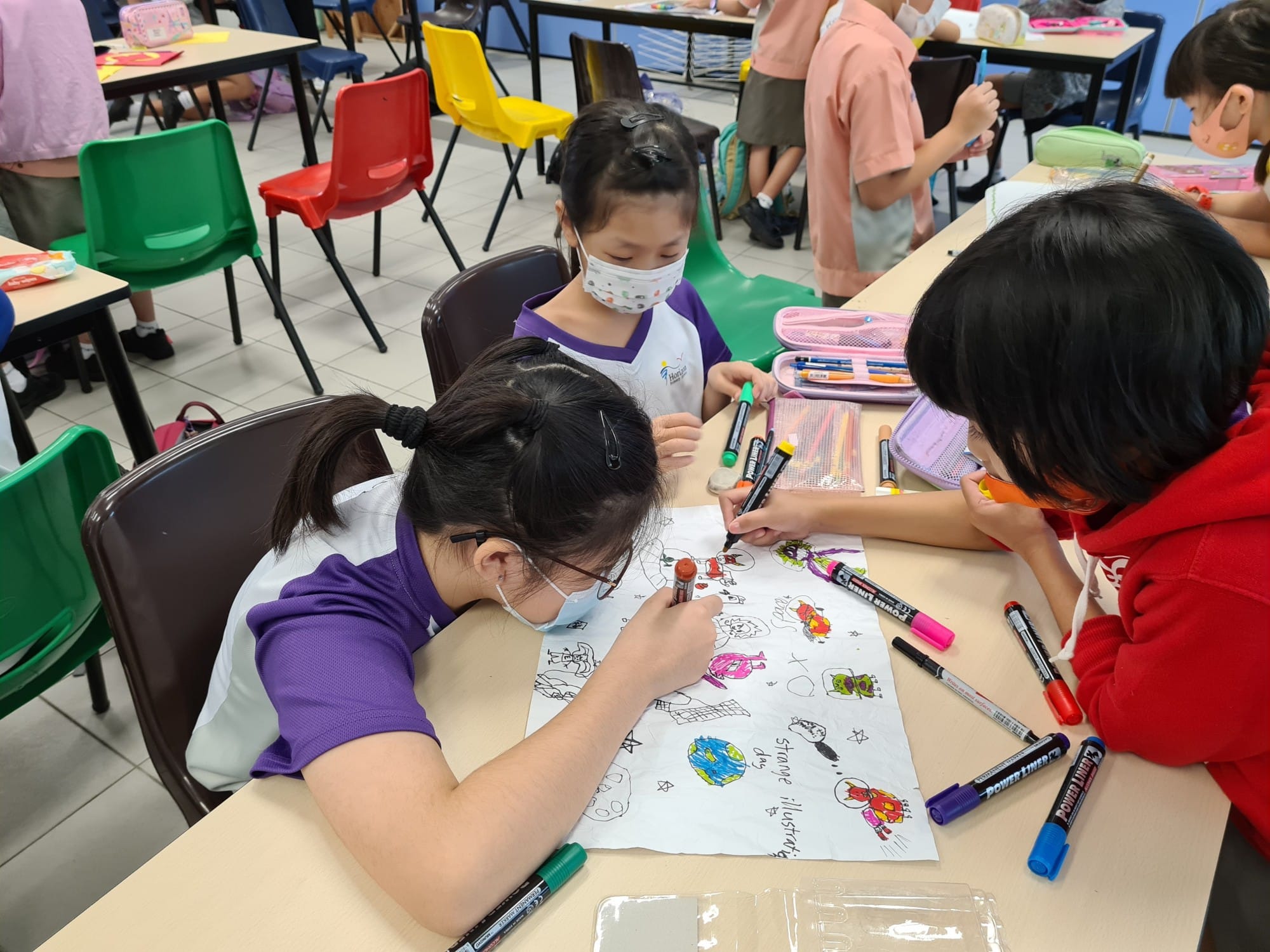 Three children wearing masks color a large drawing sheet with markers at a classroom desk.