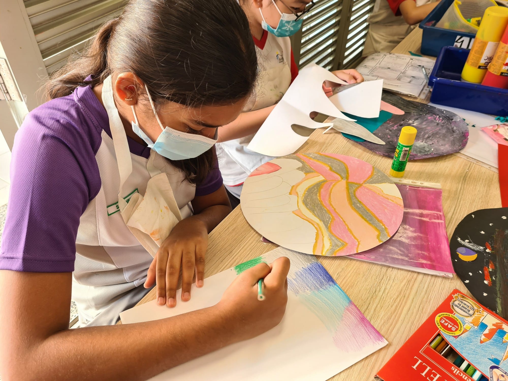 A masked student in an apron colors paper with crayons at an art table covered with colorful artwork.