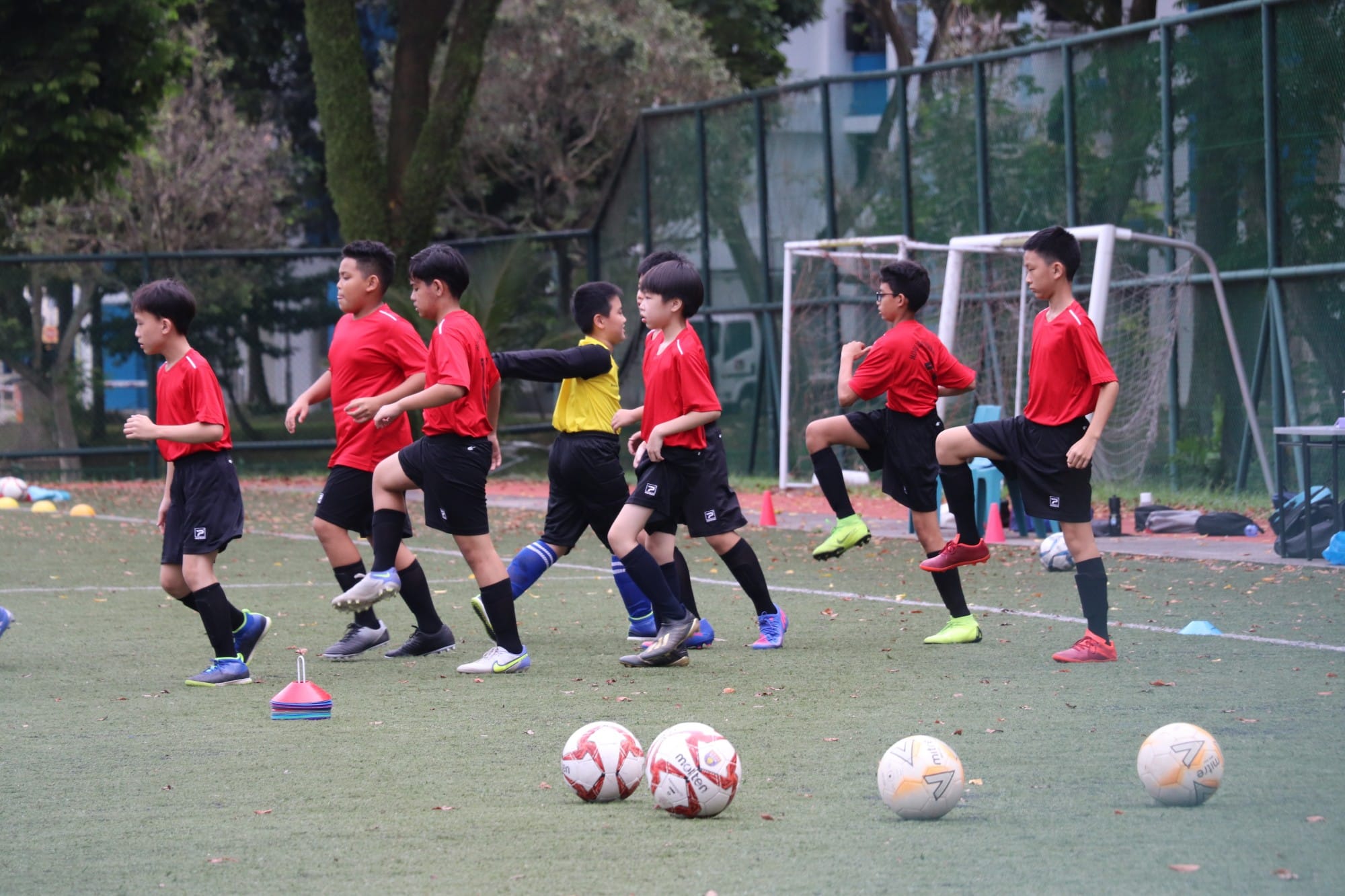 Young boys in red uniforms doing high-knee drills on a grass football field with several balls and cones nearby.