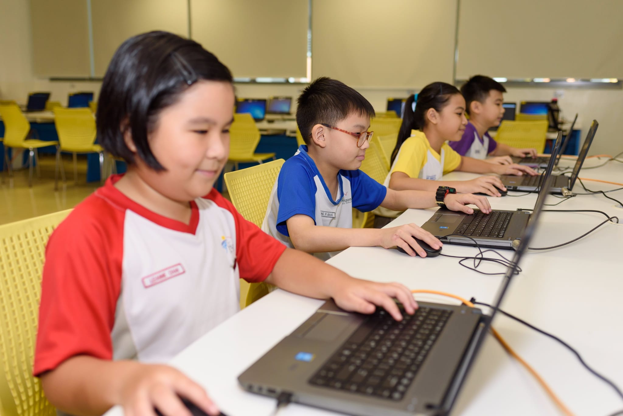 Four children in school uniforms sit in a row using laptops at a long white table in a computer lab.