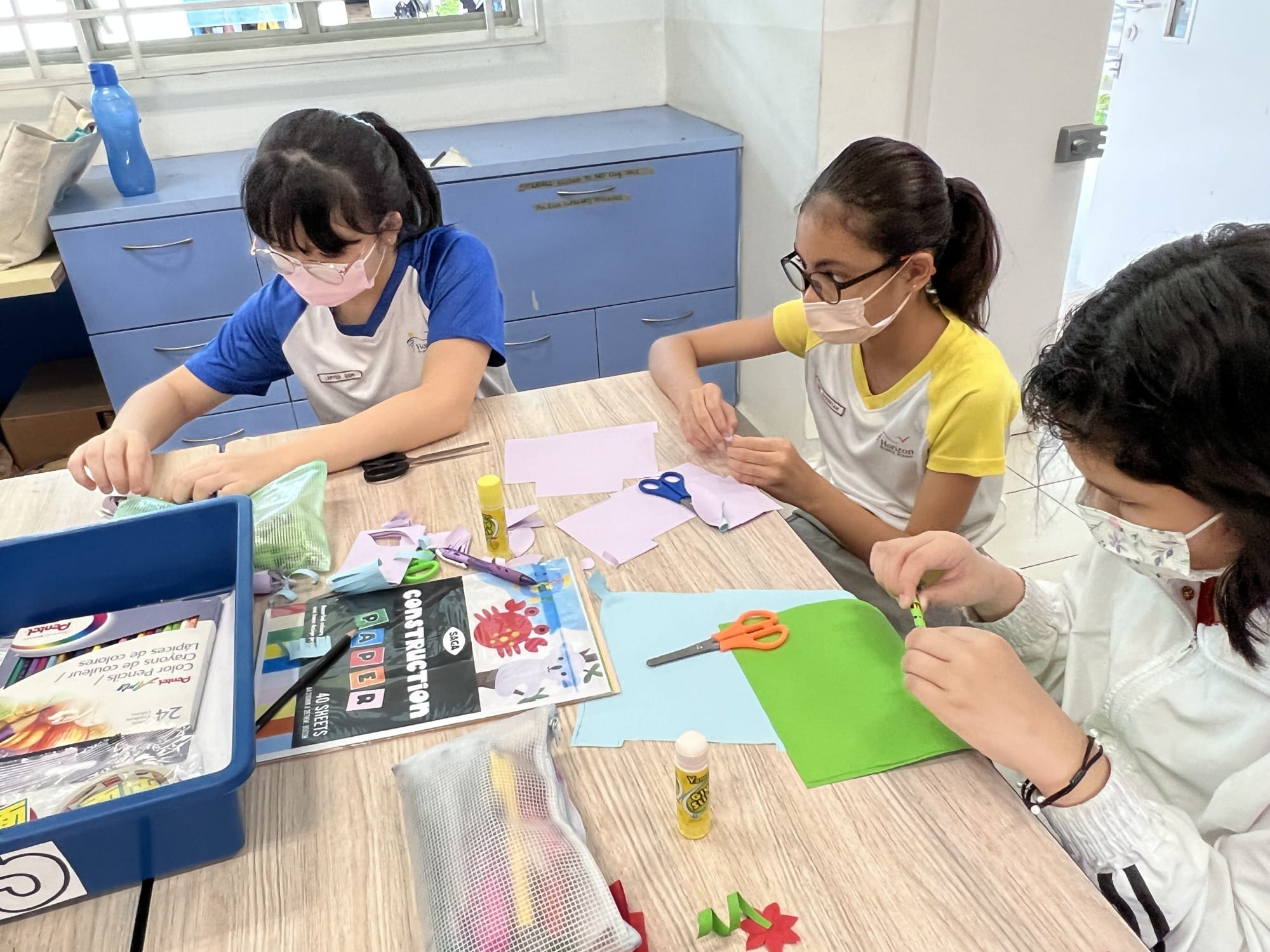Three masked girls sit at a table doing a paper craft activity with scissors, glue sticks, and colored paper.