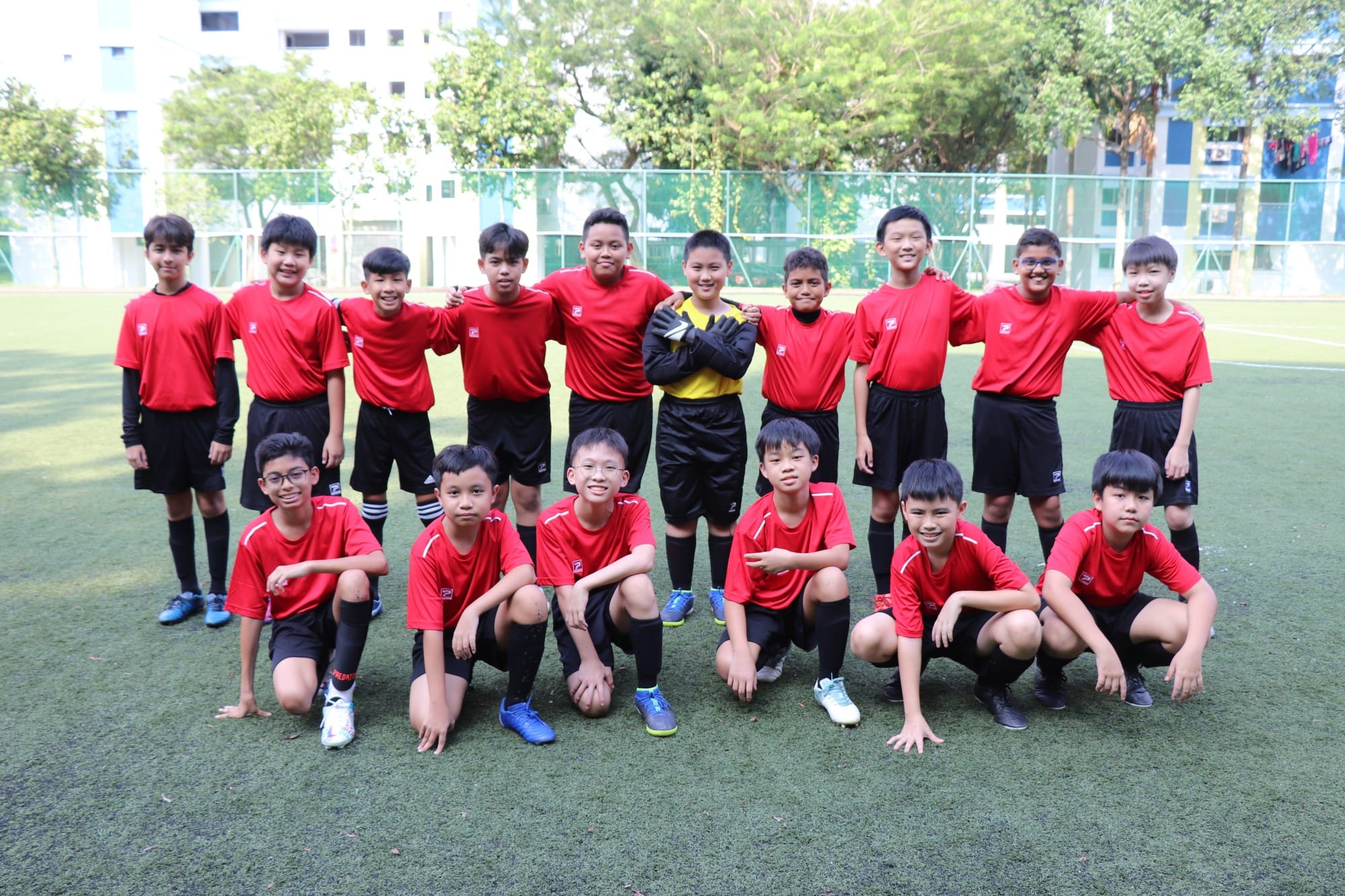 A youth football team of boys in red jerseys posing together on a green turf field, with one goalkeeper in yellow.