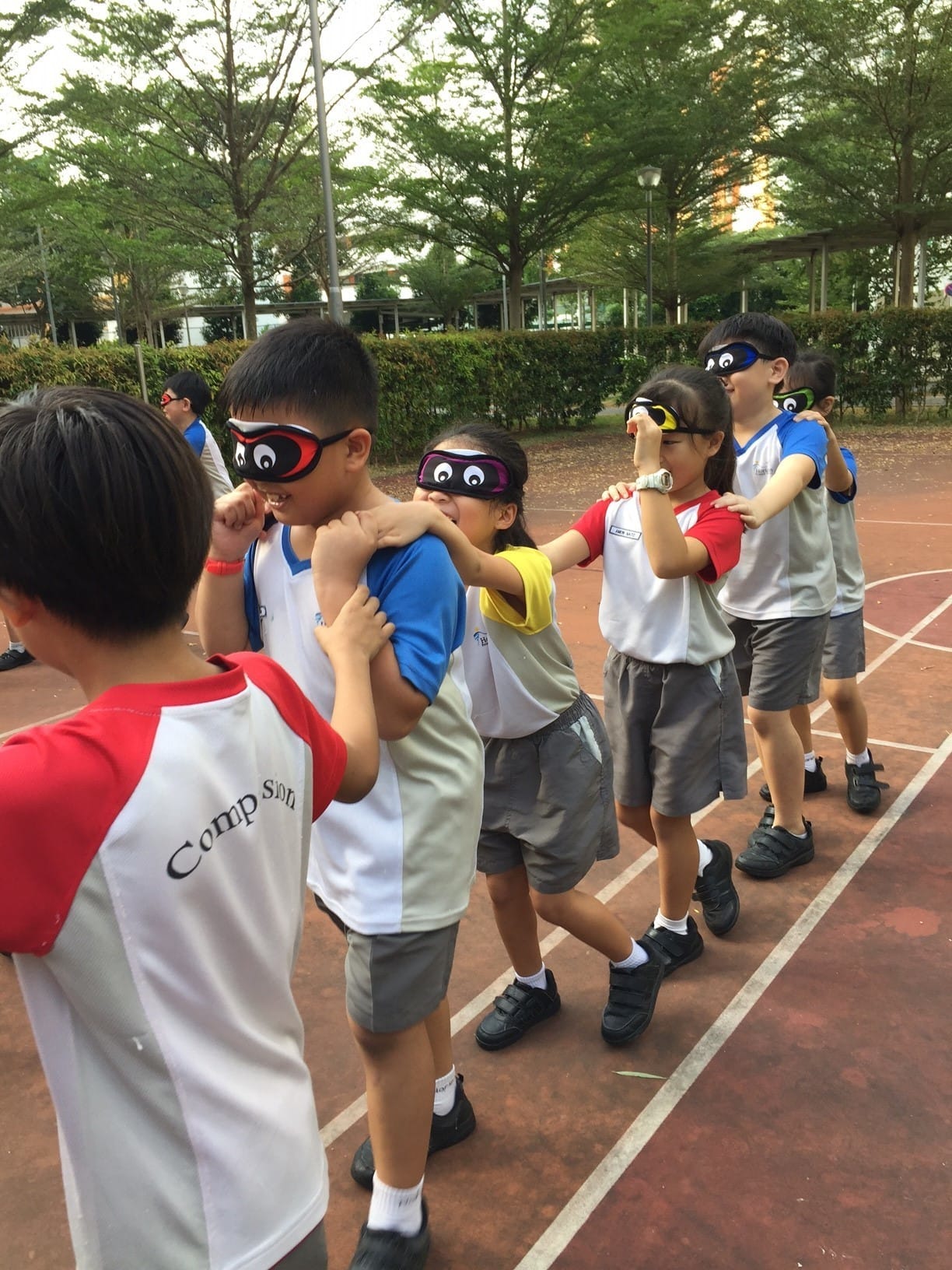 Children wearing novelty eye masks form a chain on an outdoor sports track, each placing hands on the shoulders ahead.