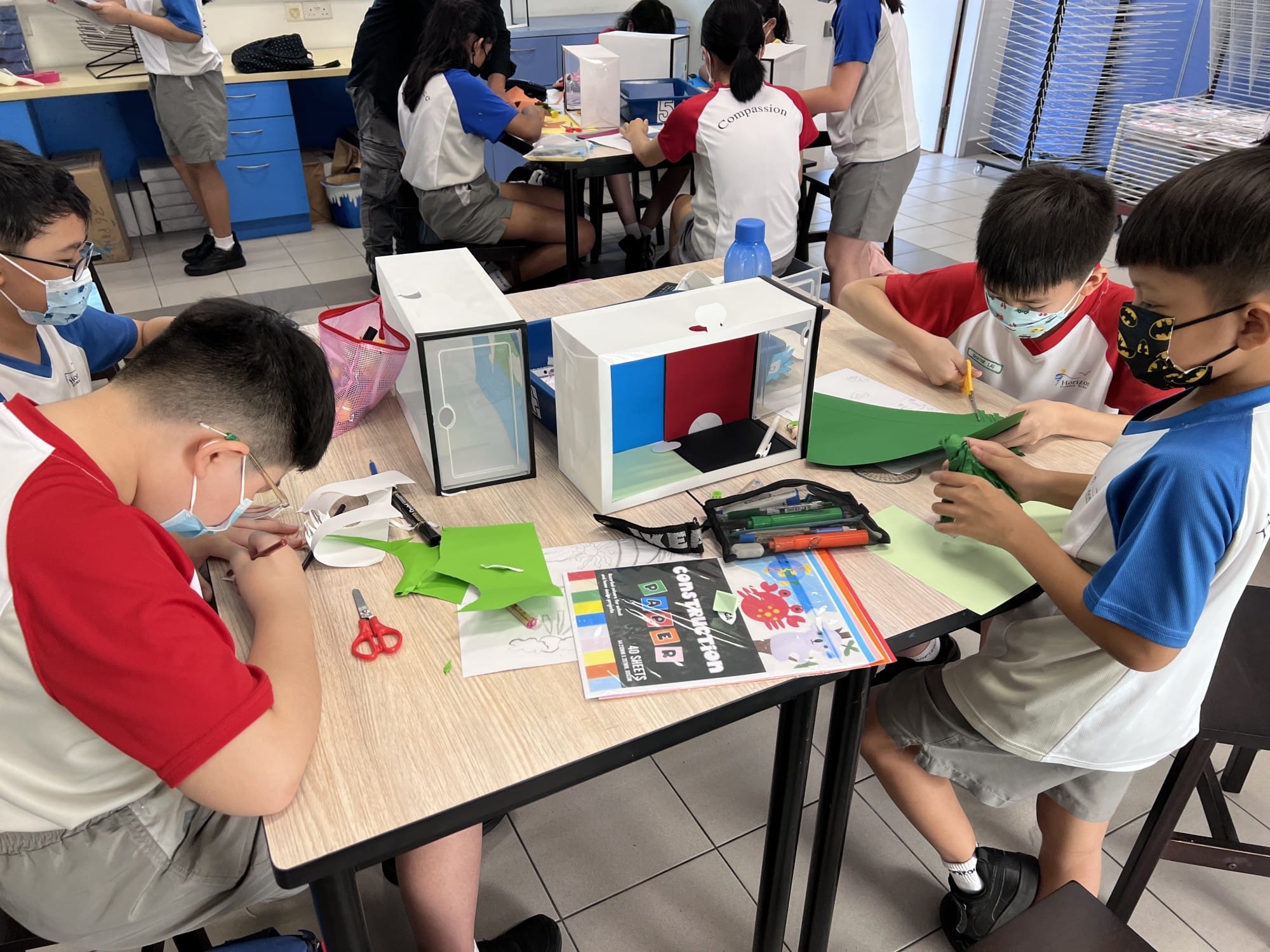 Students wearing masks do a craft activity with green construction paper, scissors, and a small box model at a classroom table.