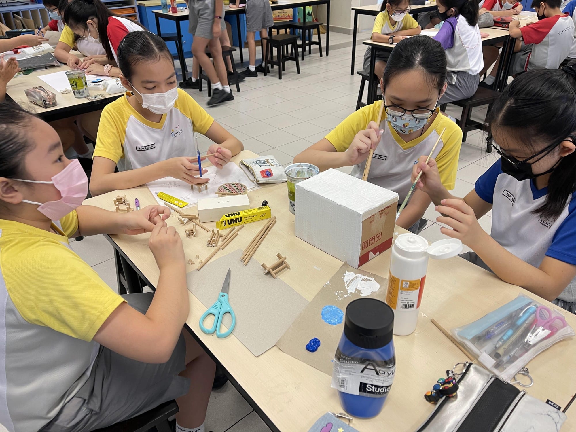 Four masked students in school uniforms work on a craft project with wood sticks, paint, glue, and a cardboard box.