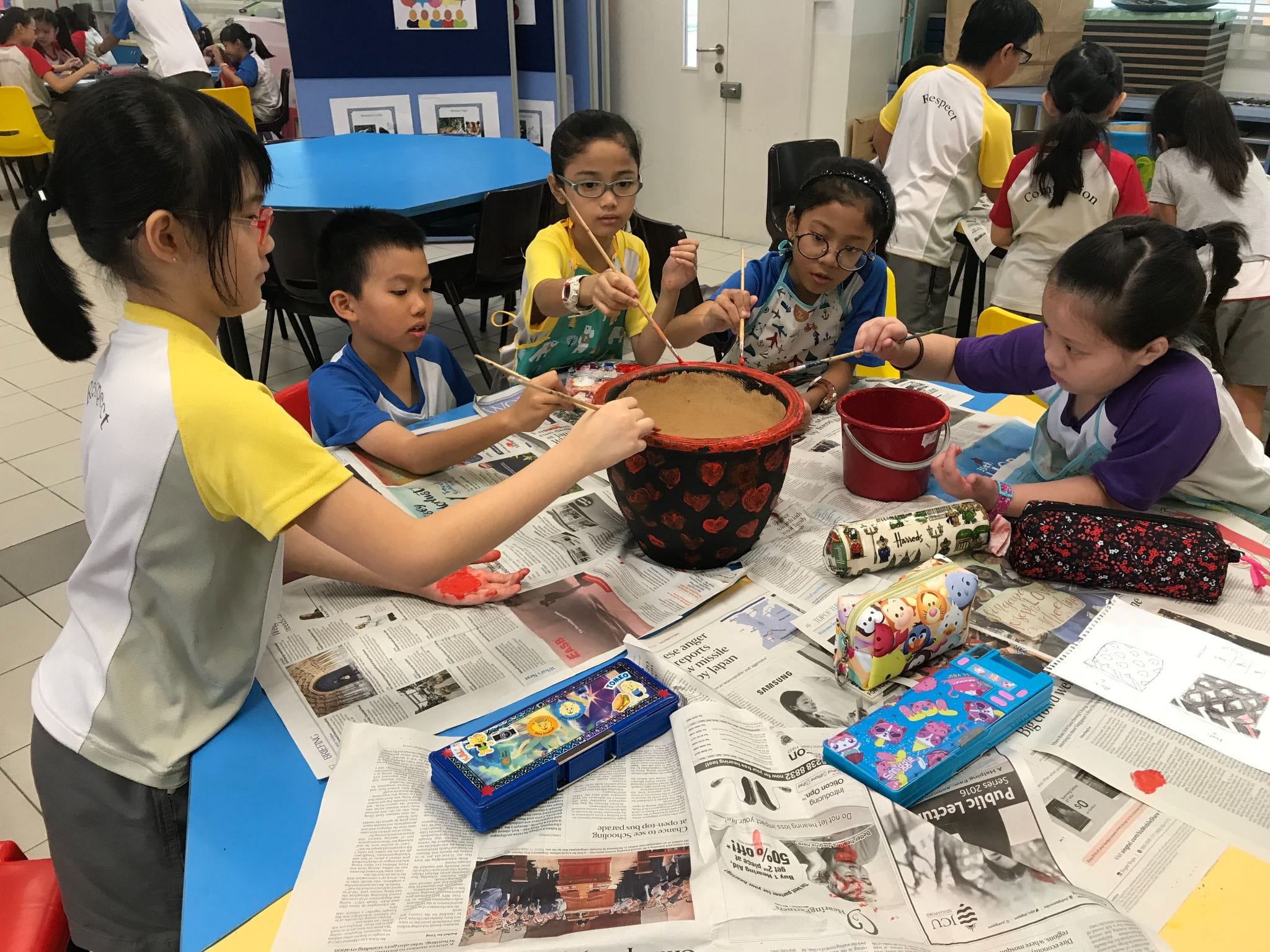 Children painting a large pot with brushes at a newspaper-covered table in a classroom setting.