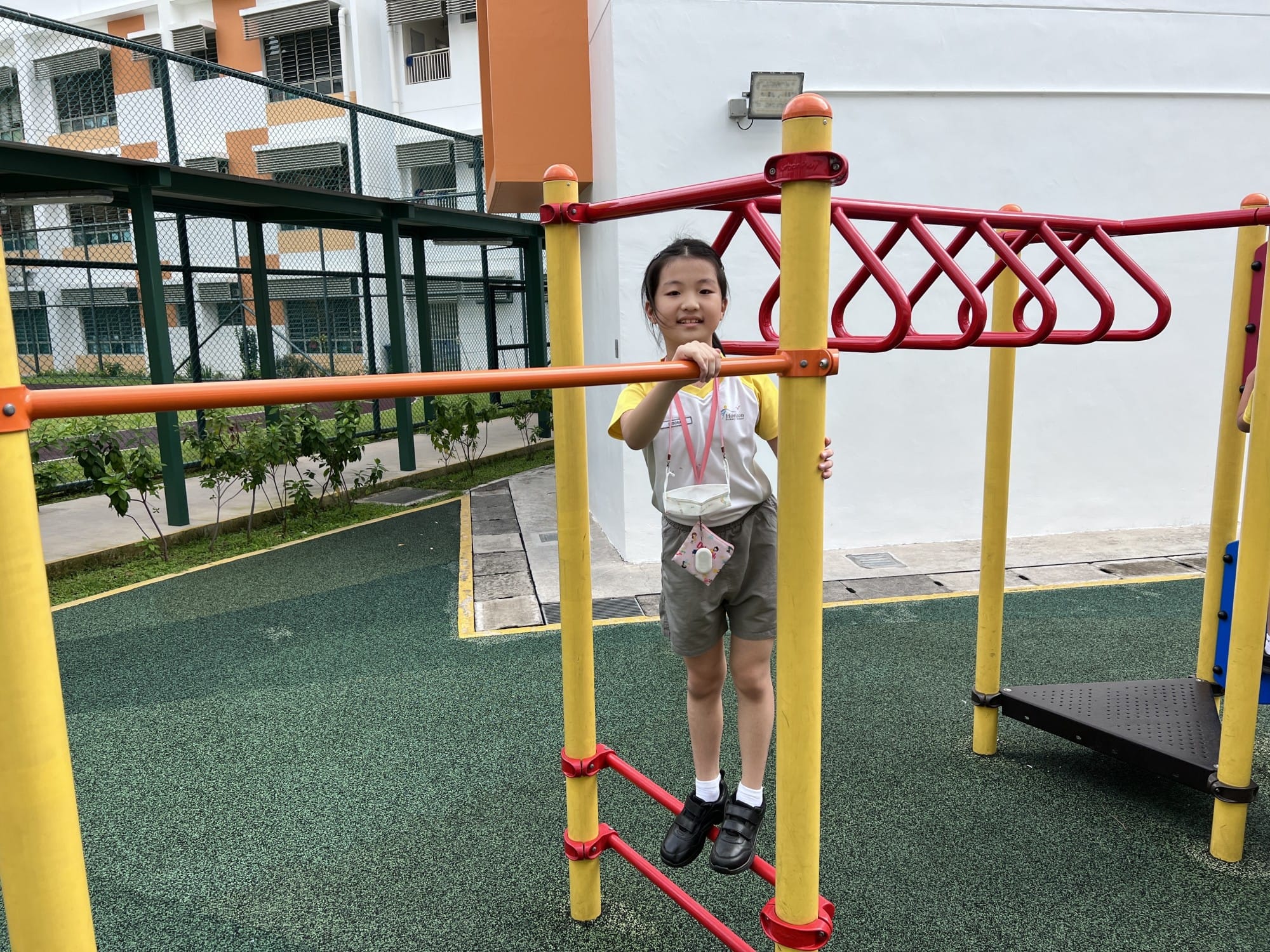 A smiling young girl in a yellow shirt holds onto yellow and red playground bars on a green rubber surface.