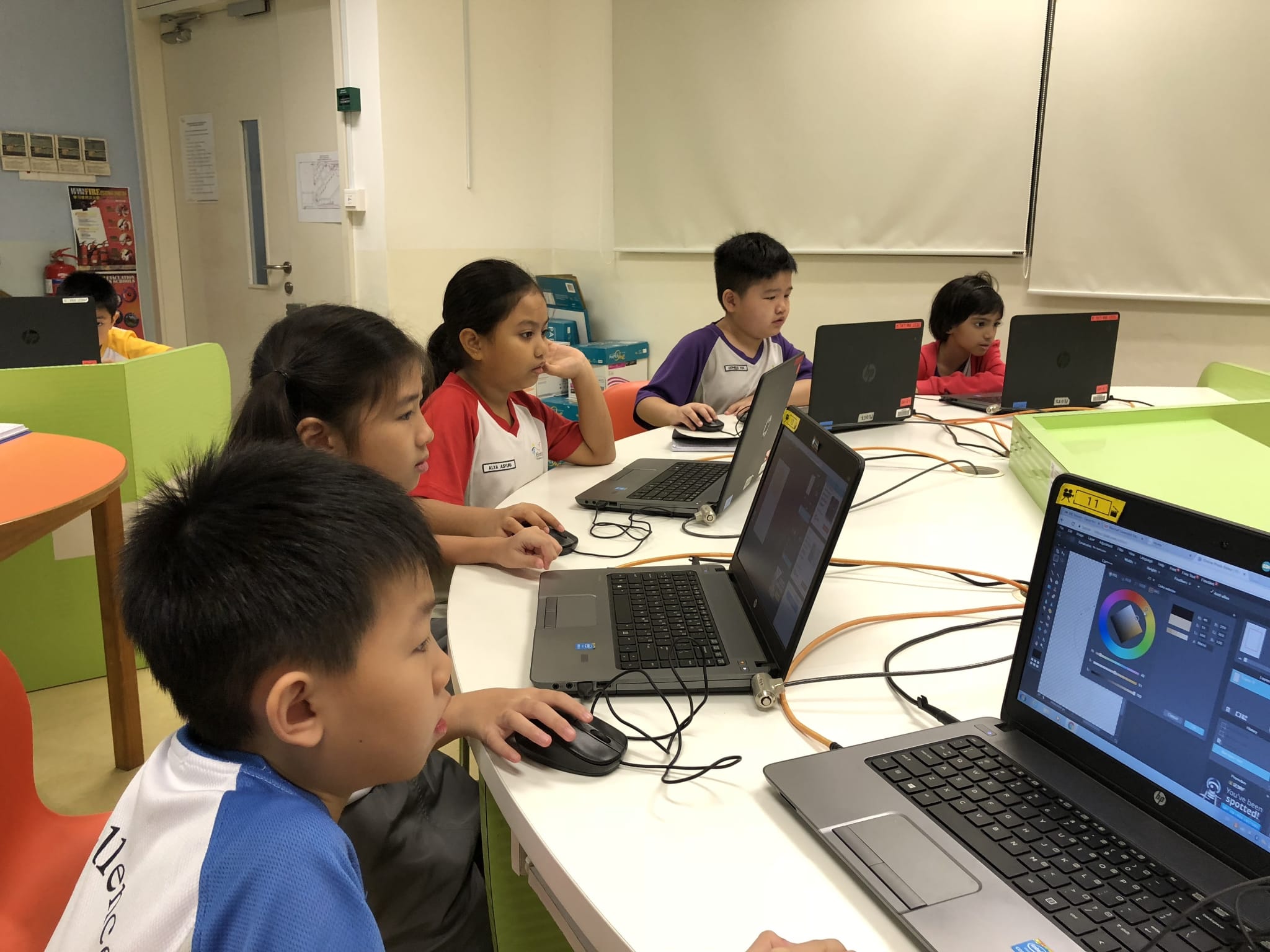 Several young students sit at a white table using HP laptops with mice in a bright classroom setting.