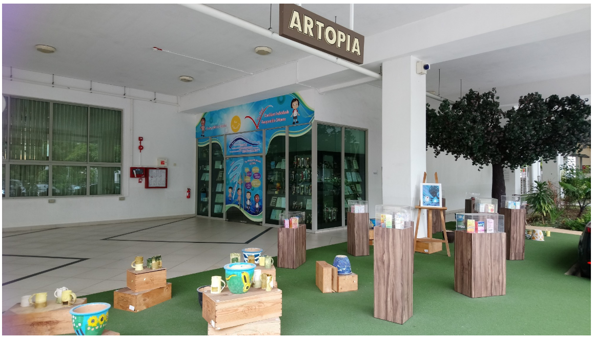 Artopia exhibition space with ceramic mugs and bowls on wooden crates and pedestals on green turf flooring.