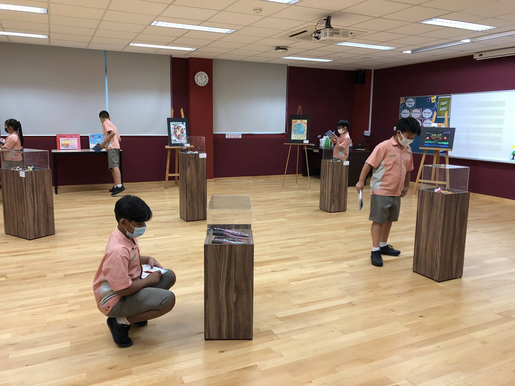 Students in pink uniforms and face masks examine display pedestals and artwork in a school exhibition hall.