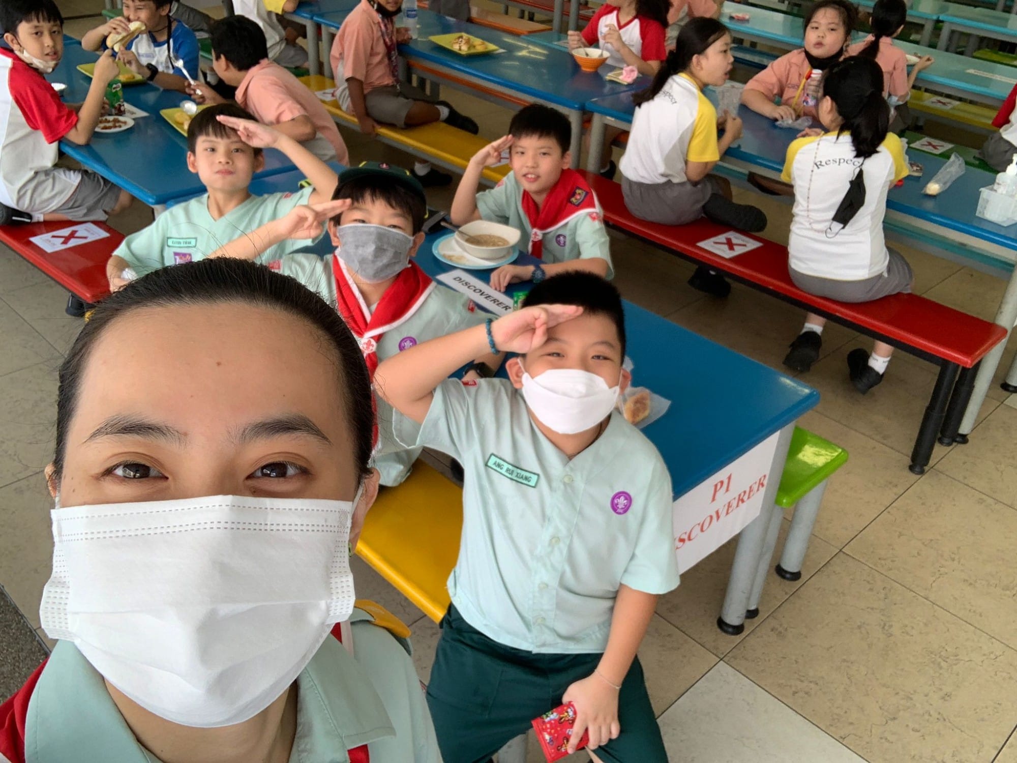 A masked adult takes a selfie with masked children in scout uniforms eating at colorful school canteen tables.