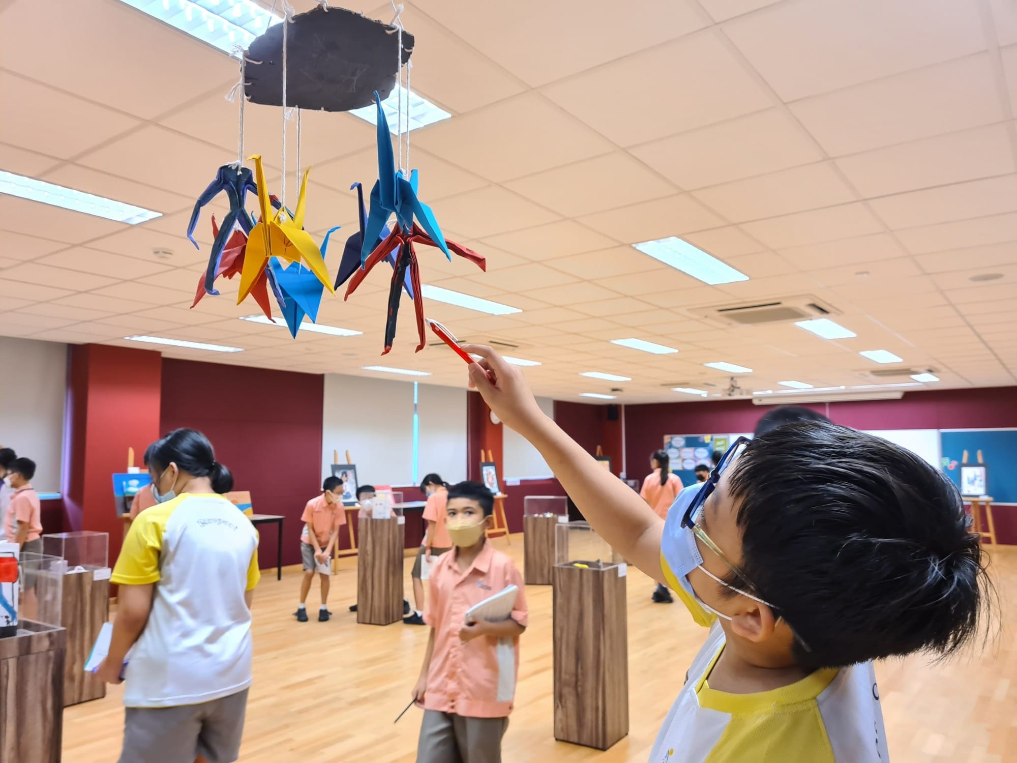 A masked child reaches up to touch colorful hanging origami cranes in a school exhibition hall with other children nearby.