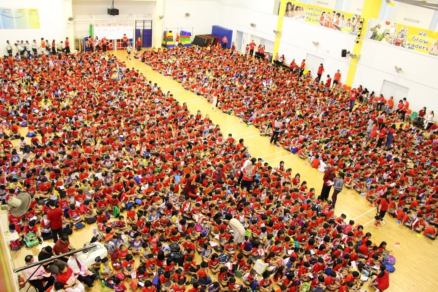 Large school hall packed with hundreds of children mostly in red shirts sitting on the floor during an assembly.
