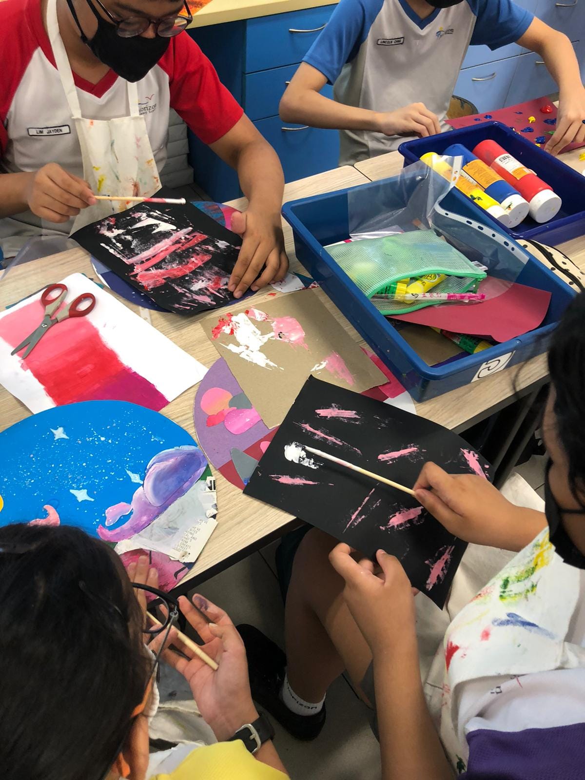 Students in aprons and masks paint colorful artwork on black and white paper at a classroom table with art supplies.