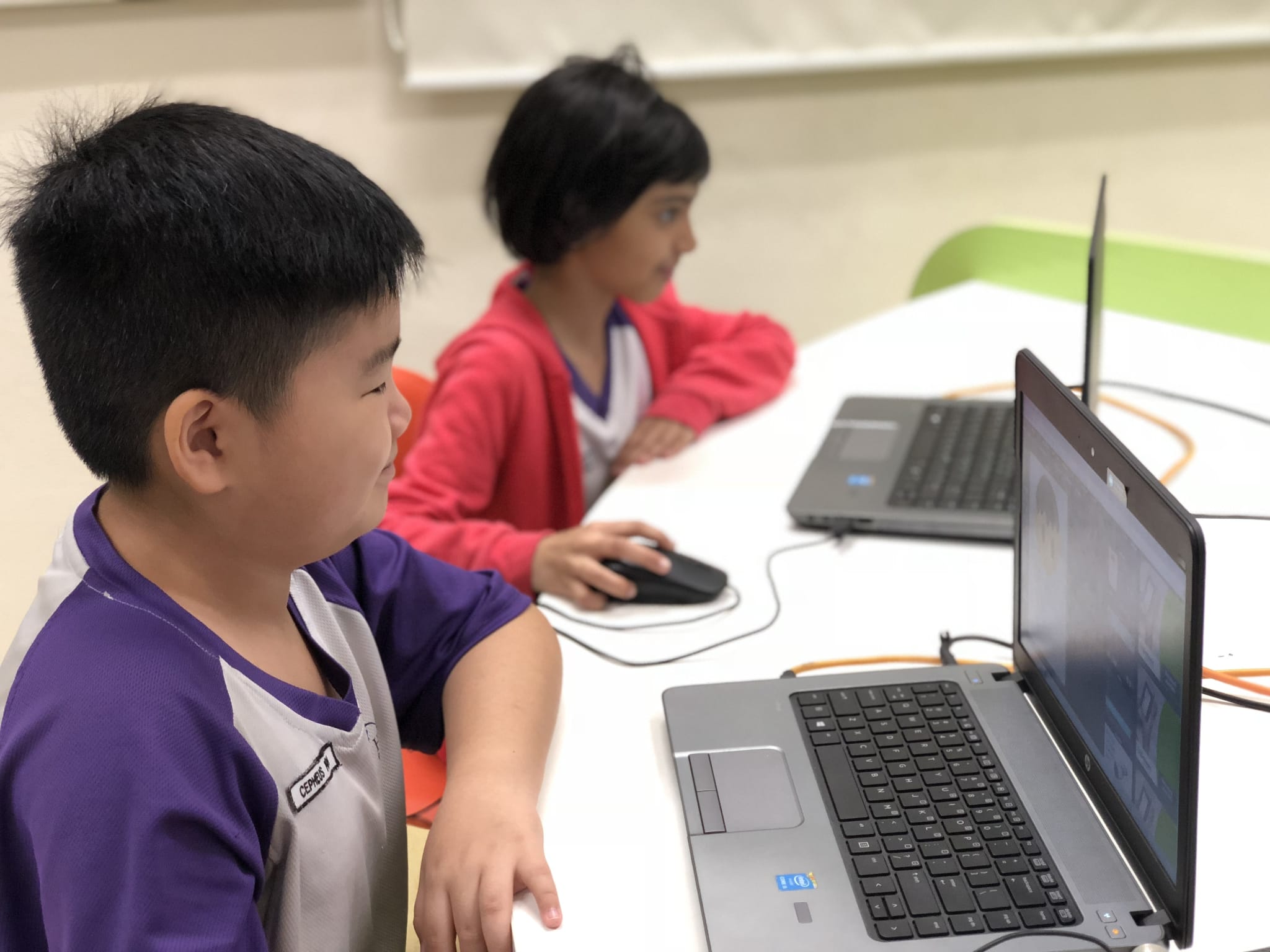 Two children sit at a white table, each using a laptop, in what appears to be a classroom setting.