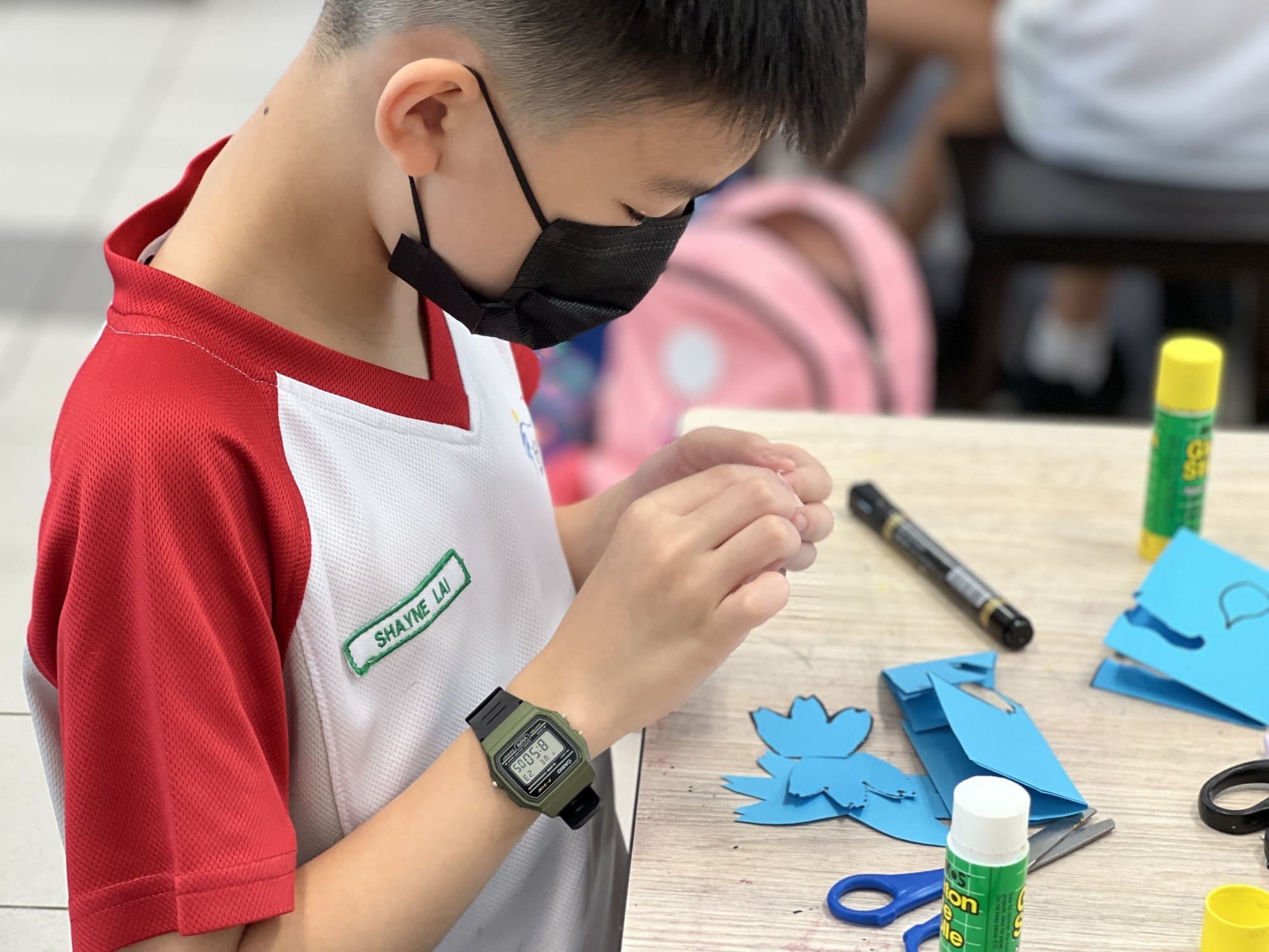 A masked boy in a school uniform works on a paper craft project with scissors and glue sticks on a desk.