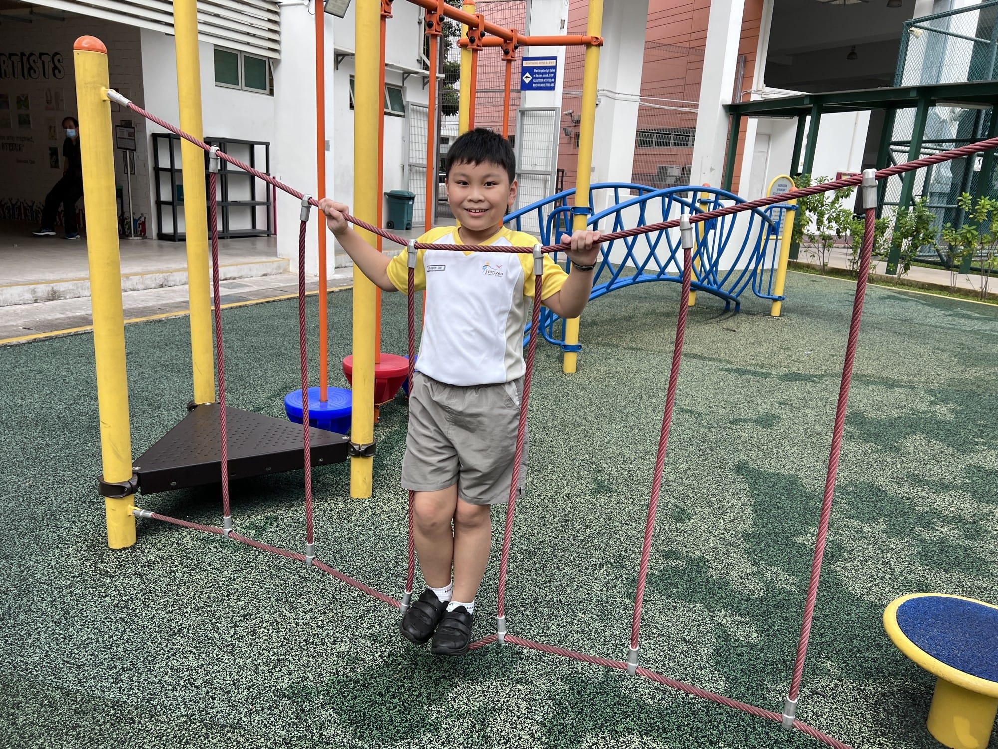 A smiling young boy in a white and yellow shirt holds rope bars on a colorful outdoor playground structure.