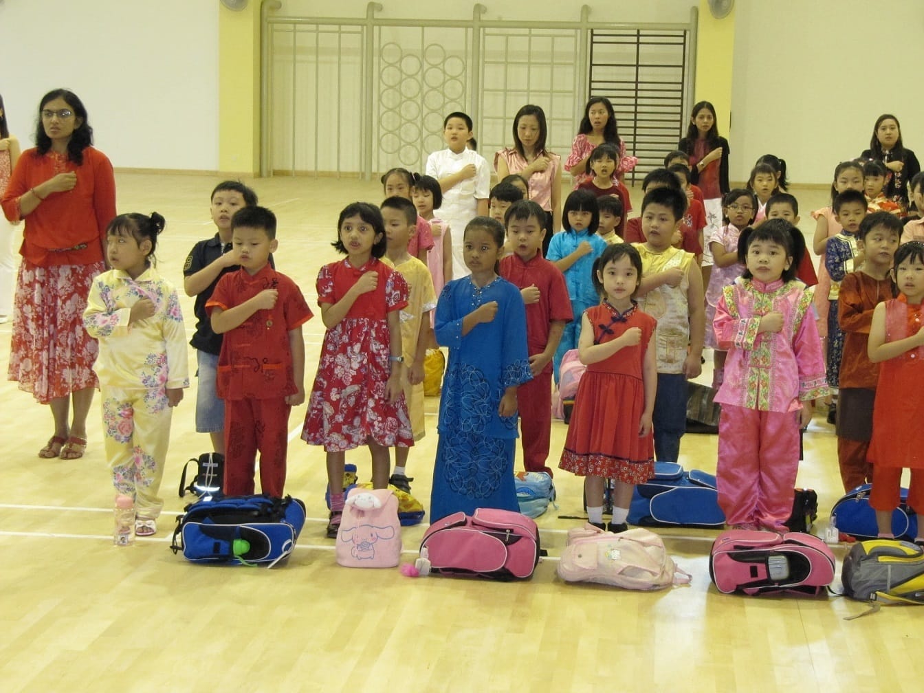Children in colorful traditional Asian outfits stand with hands on chests in a school hall, with bags on the floor.