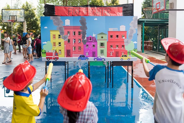 Three children in red hard hats aim water guns at a colorful firefighter-themed board game outdoors.