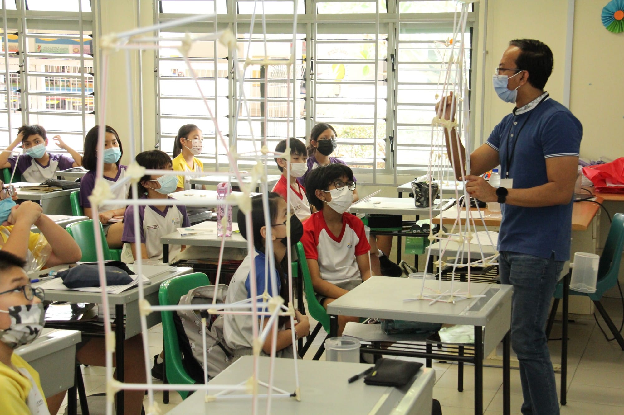 A masked teacher holds a straw tower structure while masked students sit at desks watching in a classroom.