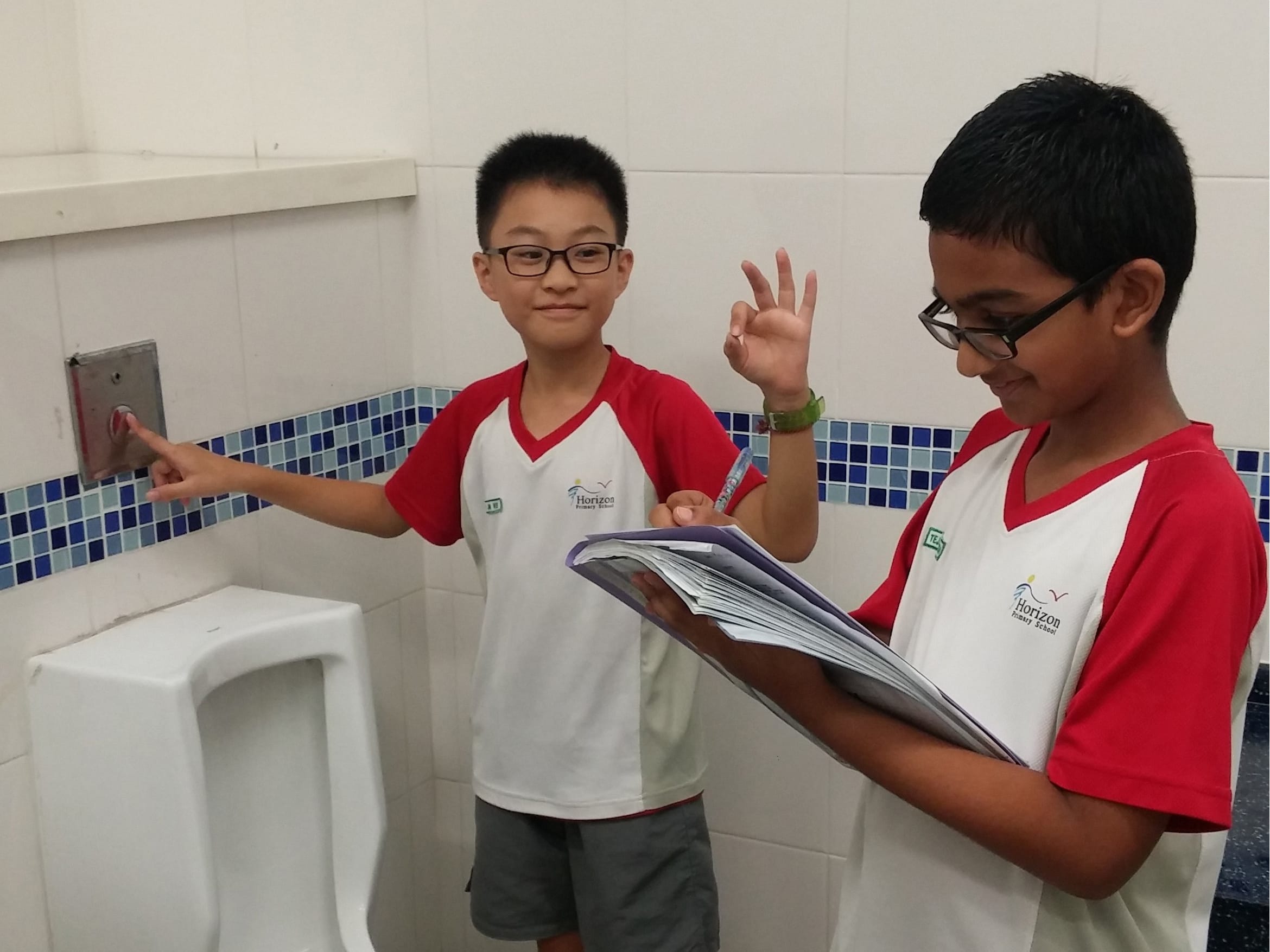 Two boys in school uniforms stand in a tiled bathroom, one pointing at a flush button while the other holds a notebook.
